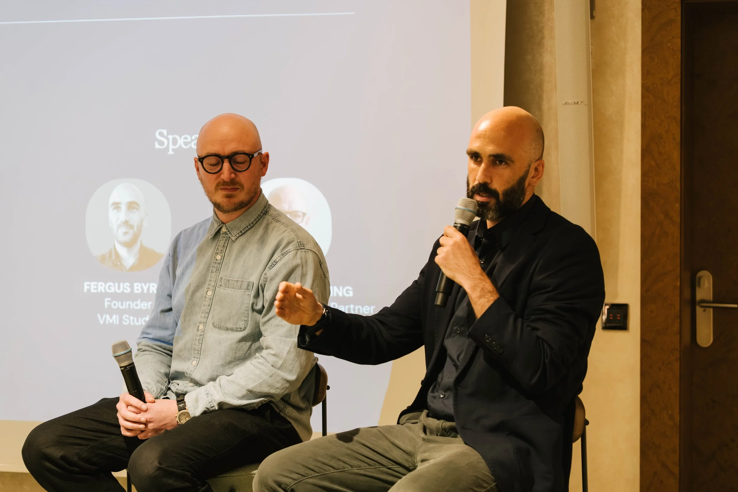 Two men sitting on chairs with microphones, participating in a panel discussion or presentation. One man is wearing glasses and a light denim shirt, holding a microphone with both hands and closed eyes. The other man has a beard and is dressed in a b