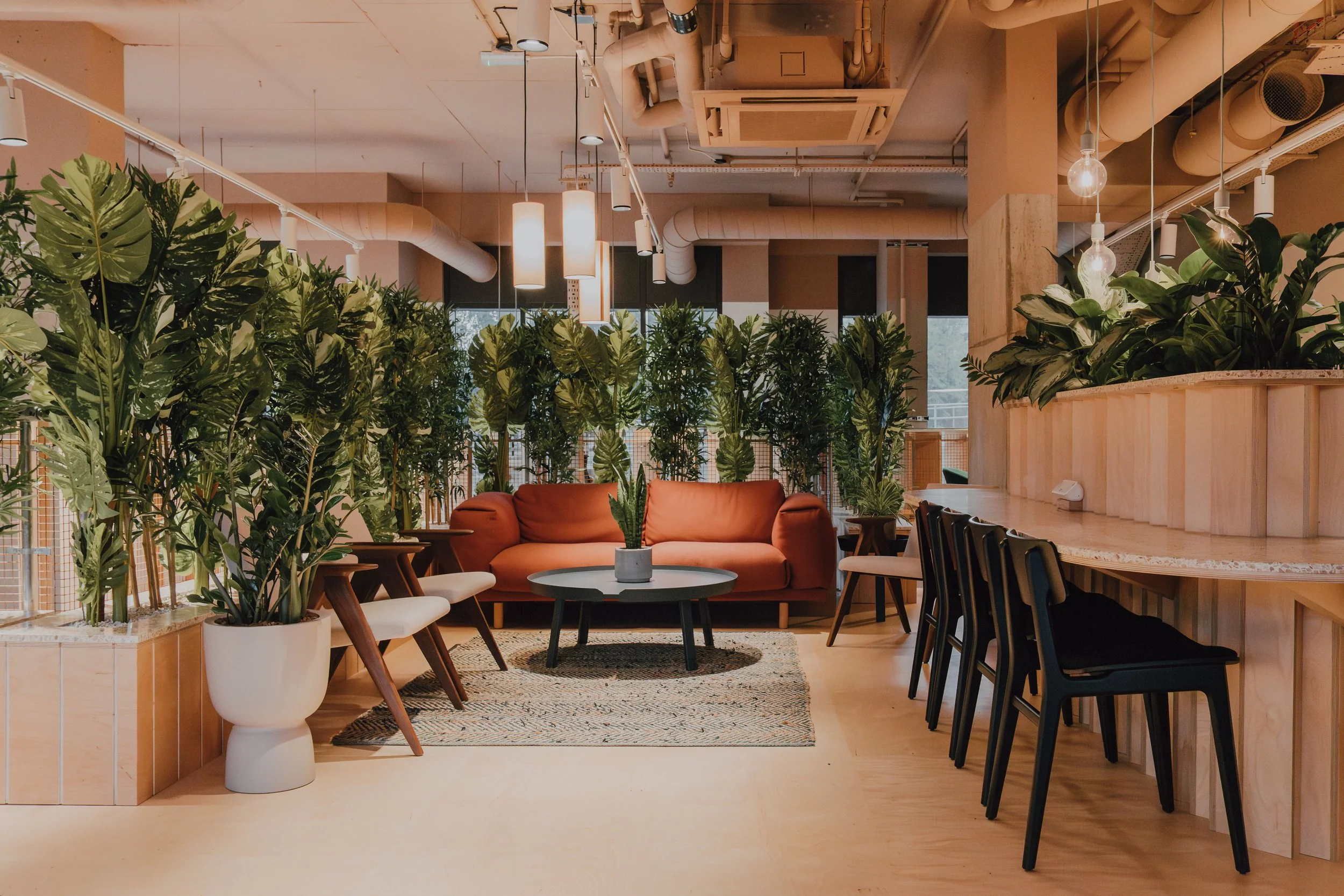 Modern indoor space with lush green plants, a cozy orange couch, and a wooden bar counter with chairs, decorated with pendant lights and large windows in the background.