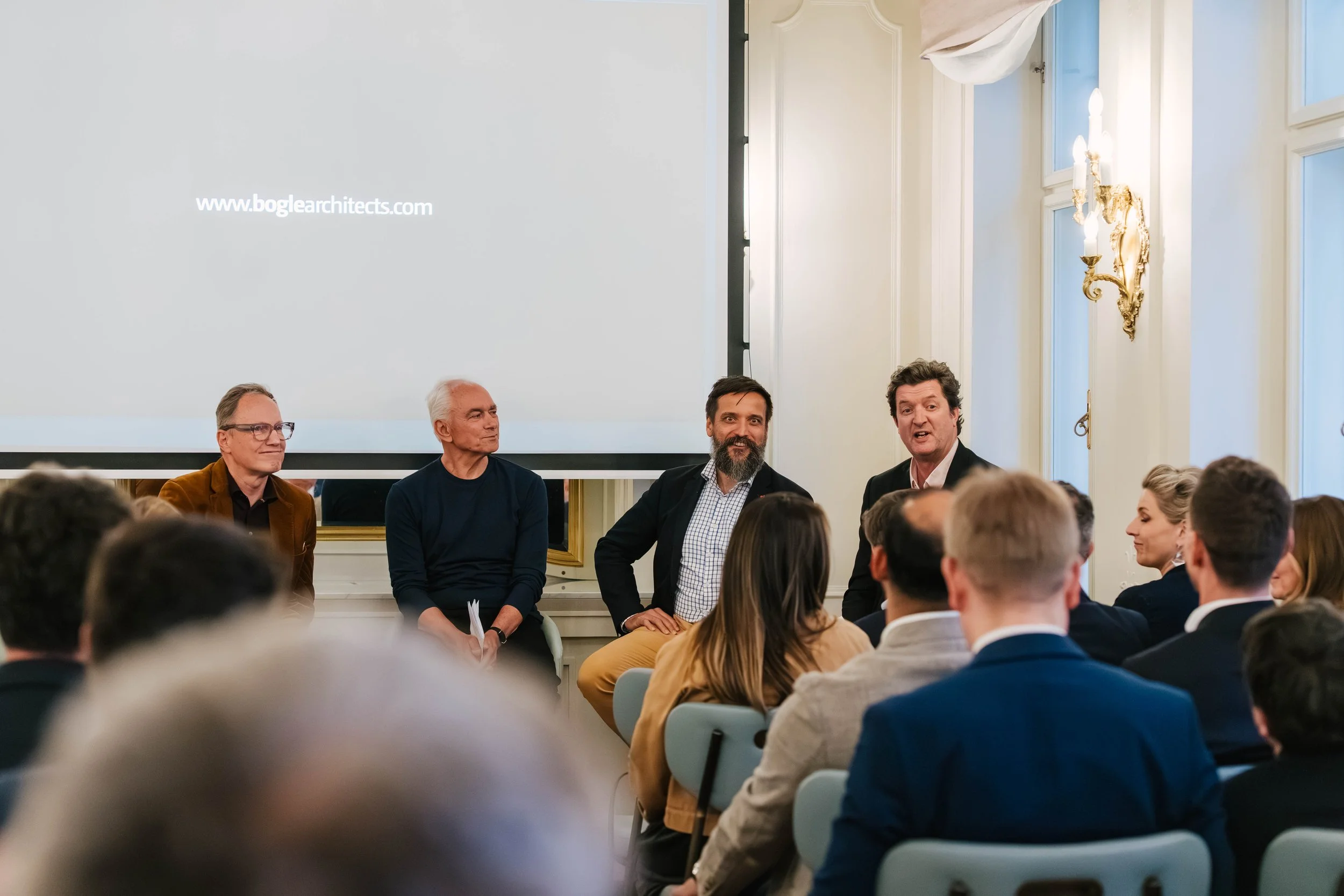 A panel of four men seated at the front of a room during a presentation or conference, facing an audience. The room has ornate white walls, large windows, and decorative gold wall sconces.