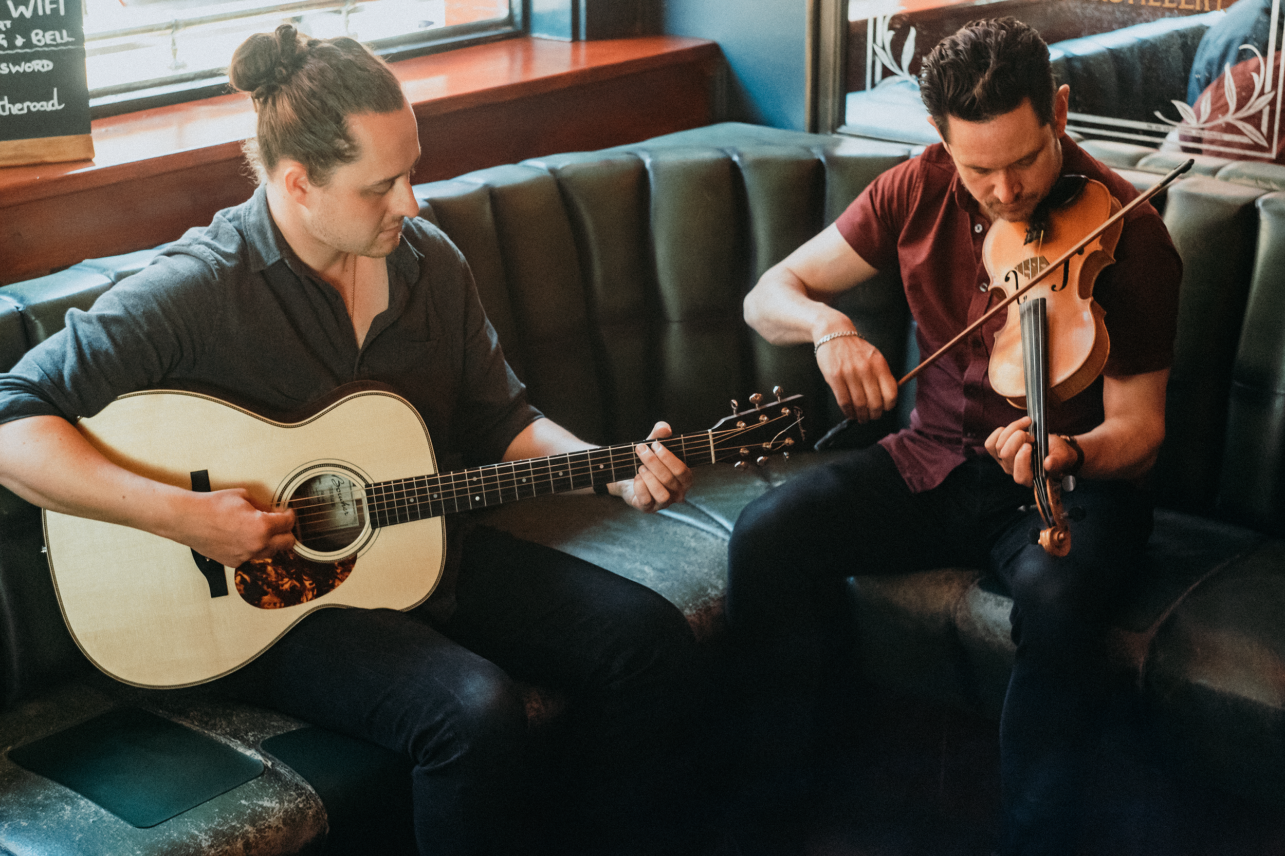 Two men sitting in a cozy cafe, playing guitars and a violin together near a window.