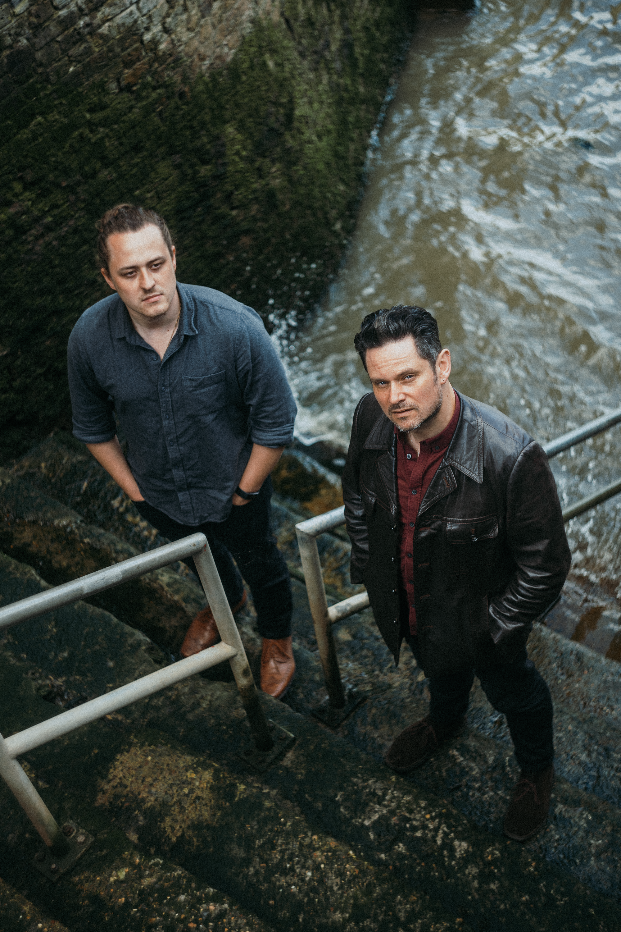 Two men standing next to a moss-covered brick wall and water, looking up at the camera. One man has dark hair and is wearing a leather jacket, the other has brown hair tied back and is wearing a blue shirt.