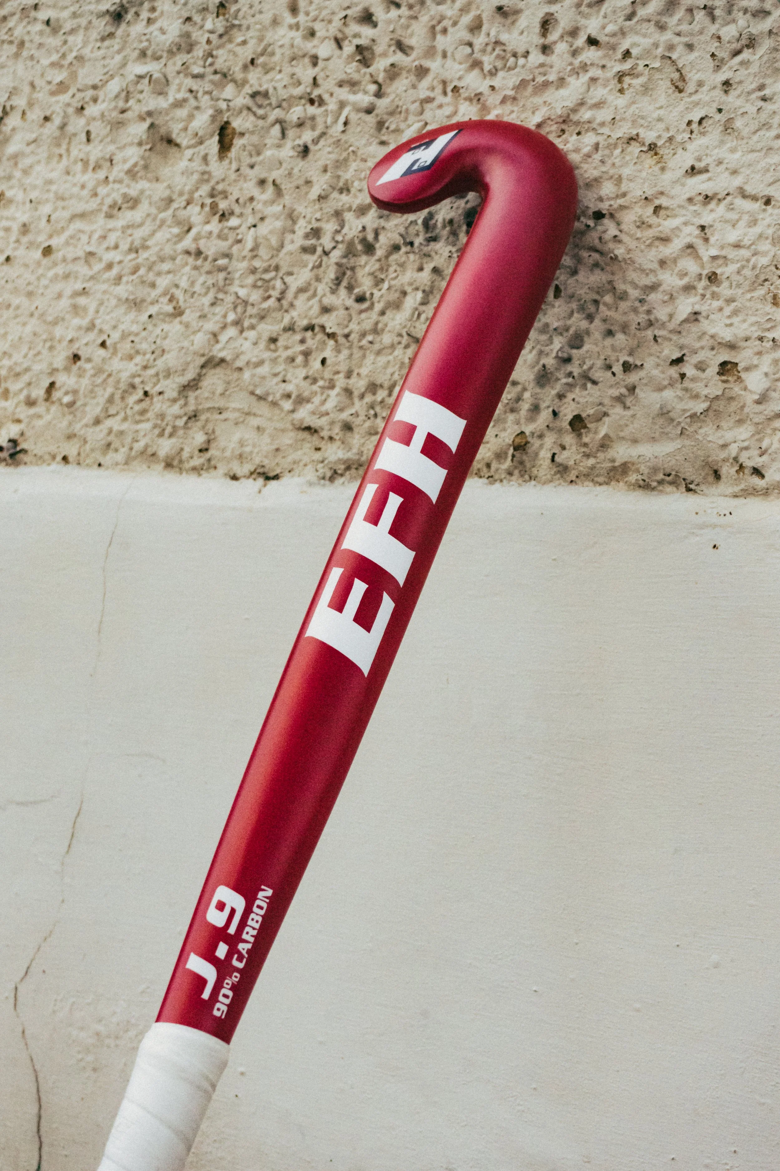 Red tennis racket with white grip, leaning against a textured beige wall.