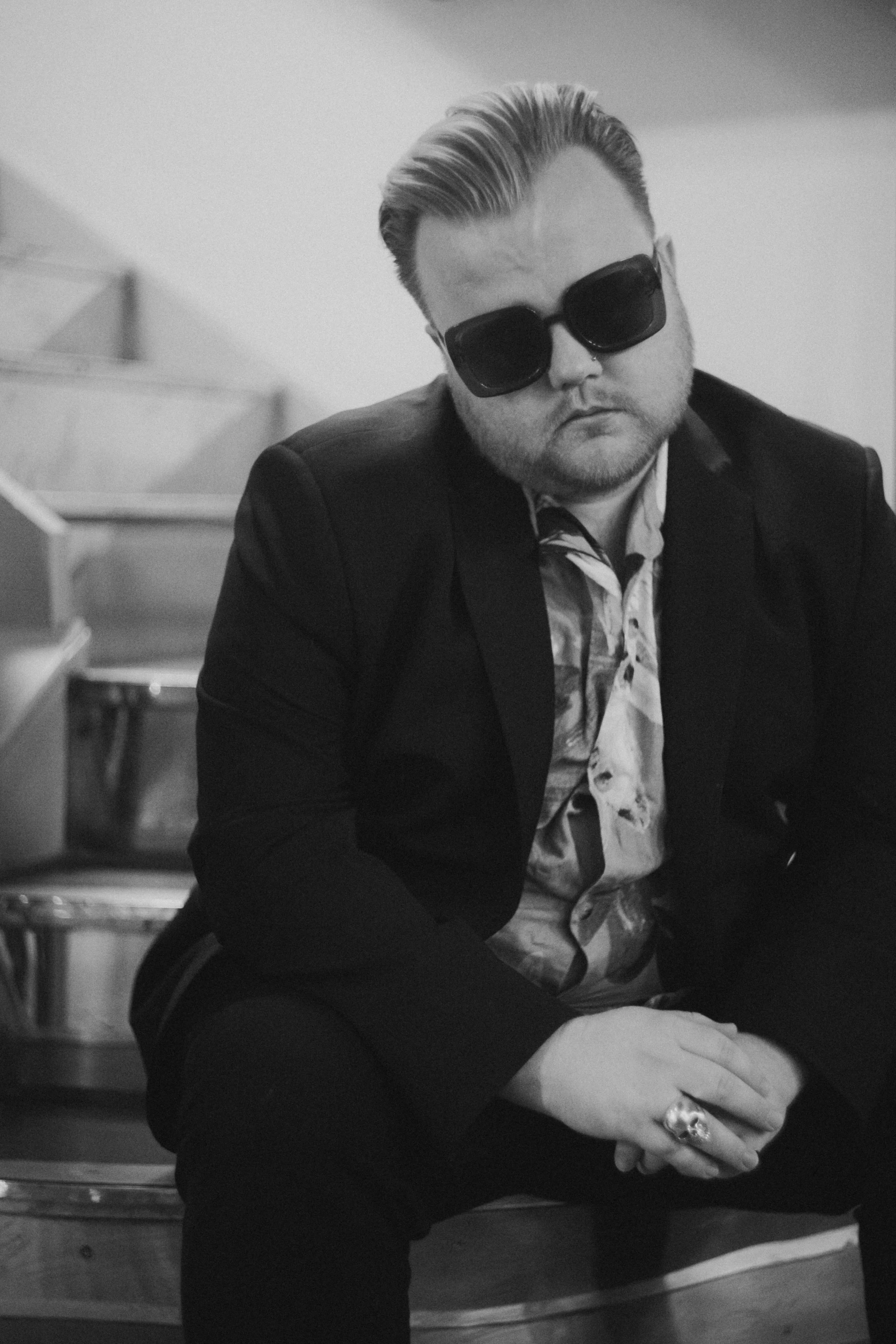 Black and white photo of a man wearing large dark sunglasses, with slicked-back hair, a nose piercing, a patterned shirt, and a jacket, seated indoors with a contemplative pose.