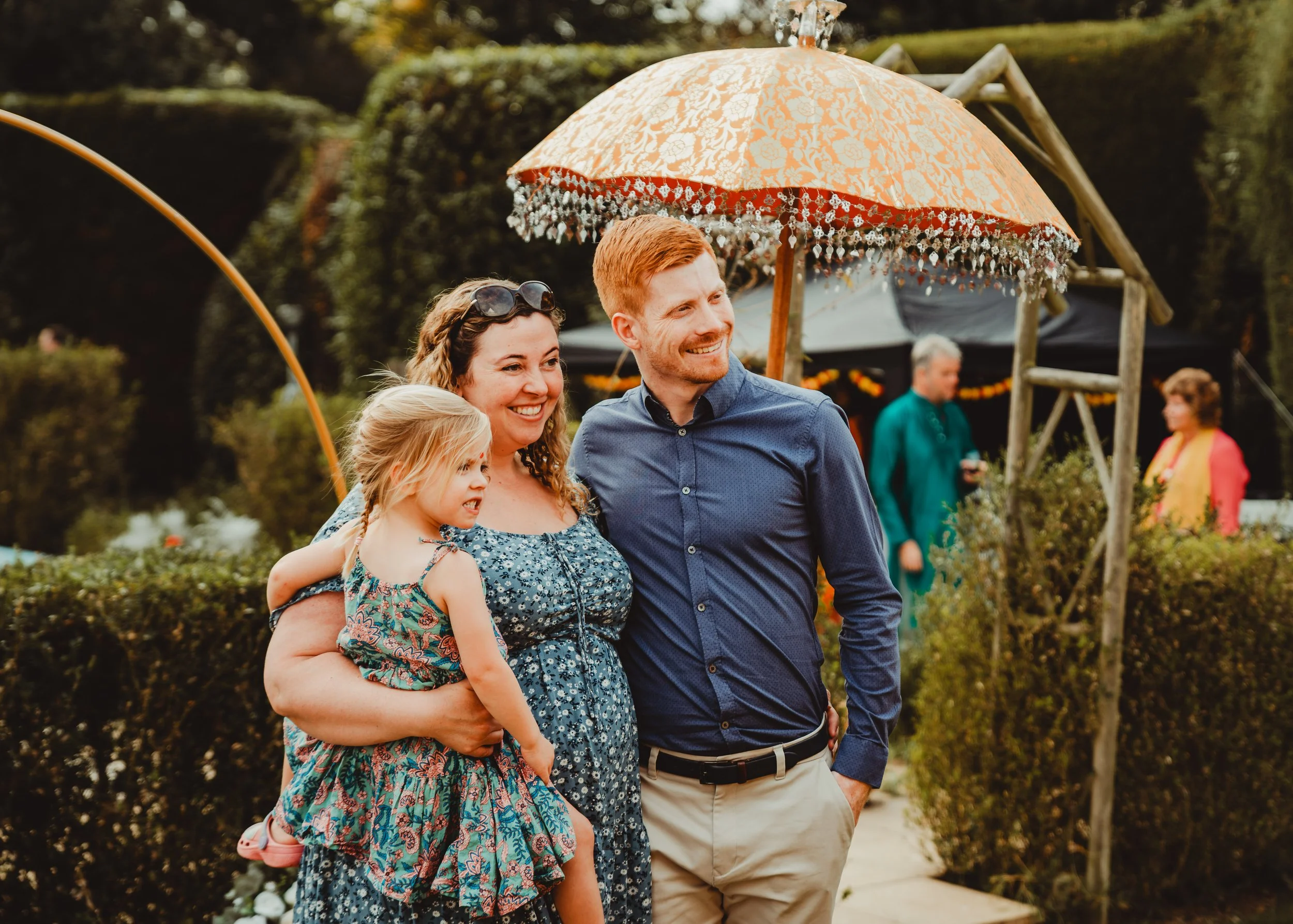 A family of four enjoying outdoors at a garden party, with a woman holding a young girl, a man smiling, all under a decorative umbrella.