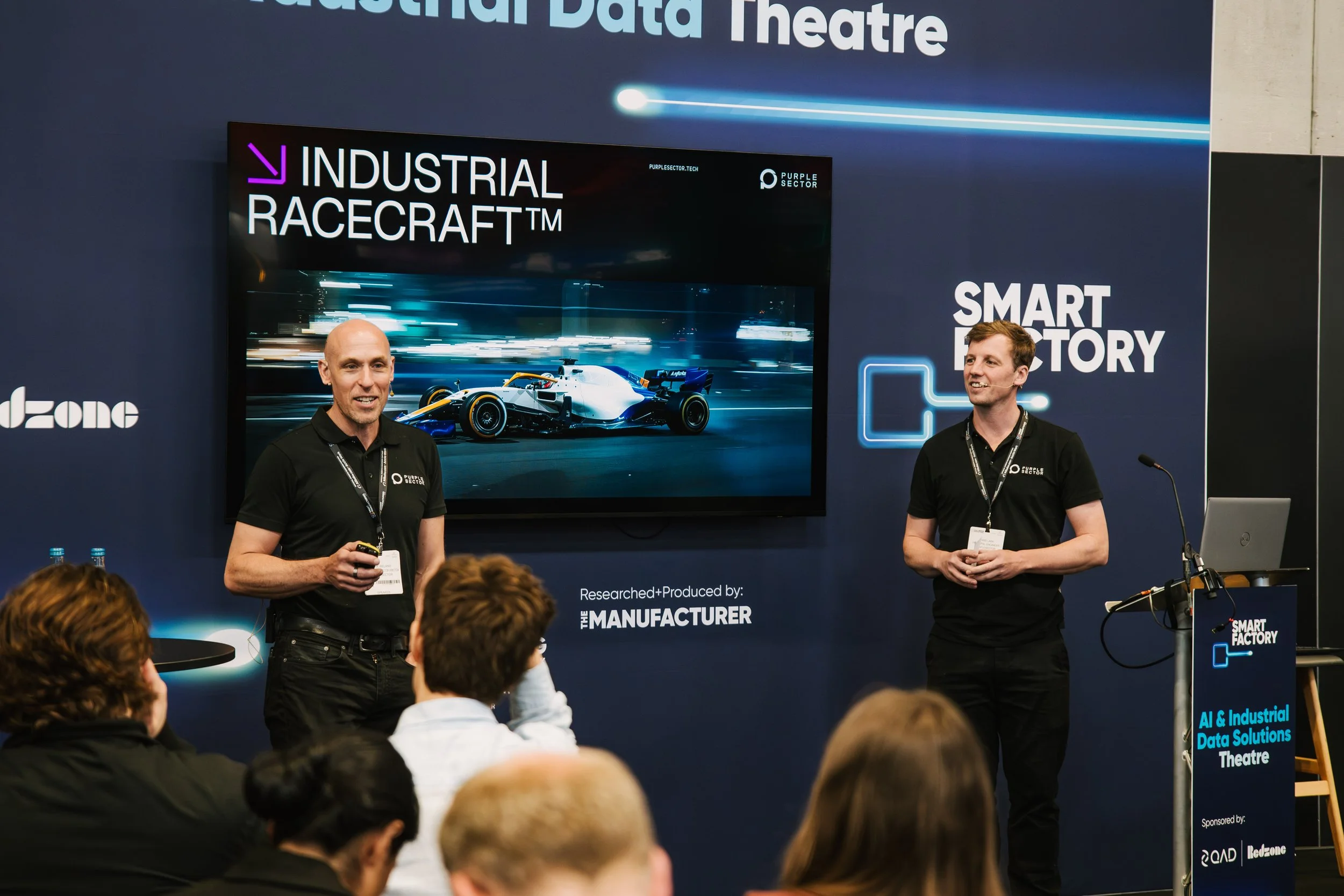 Two men standing in front of a presentation screen at the 'Industrial Data Theatre' event. The screen displays a racing car and the title 'INDUSTRIAL RACECAR™'. The audience is seated and listening to the speakers.