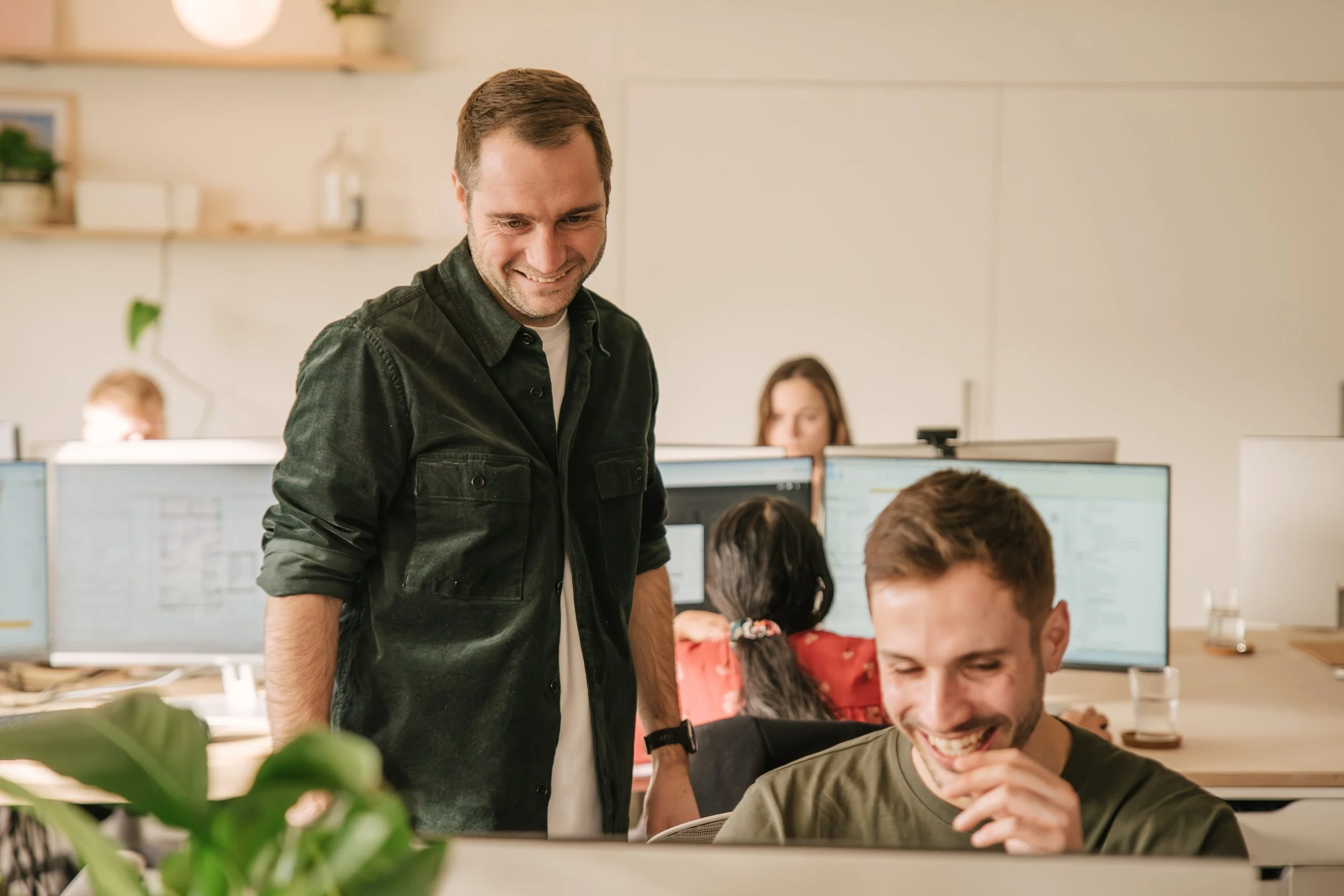 Group of people working together in an office, with a man smiling and looking down at a computer screen, surrounded by colleagues who are also engaged and smiling.