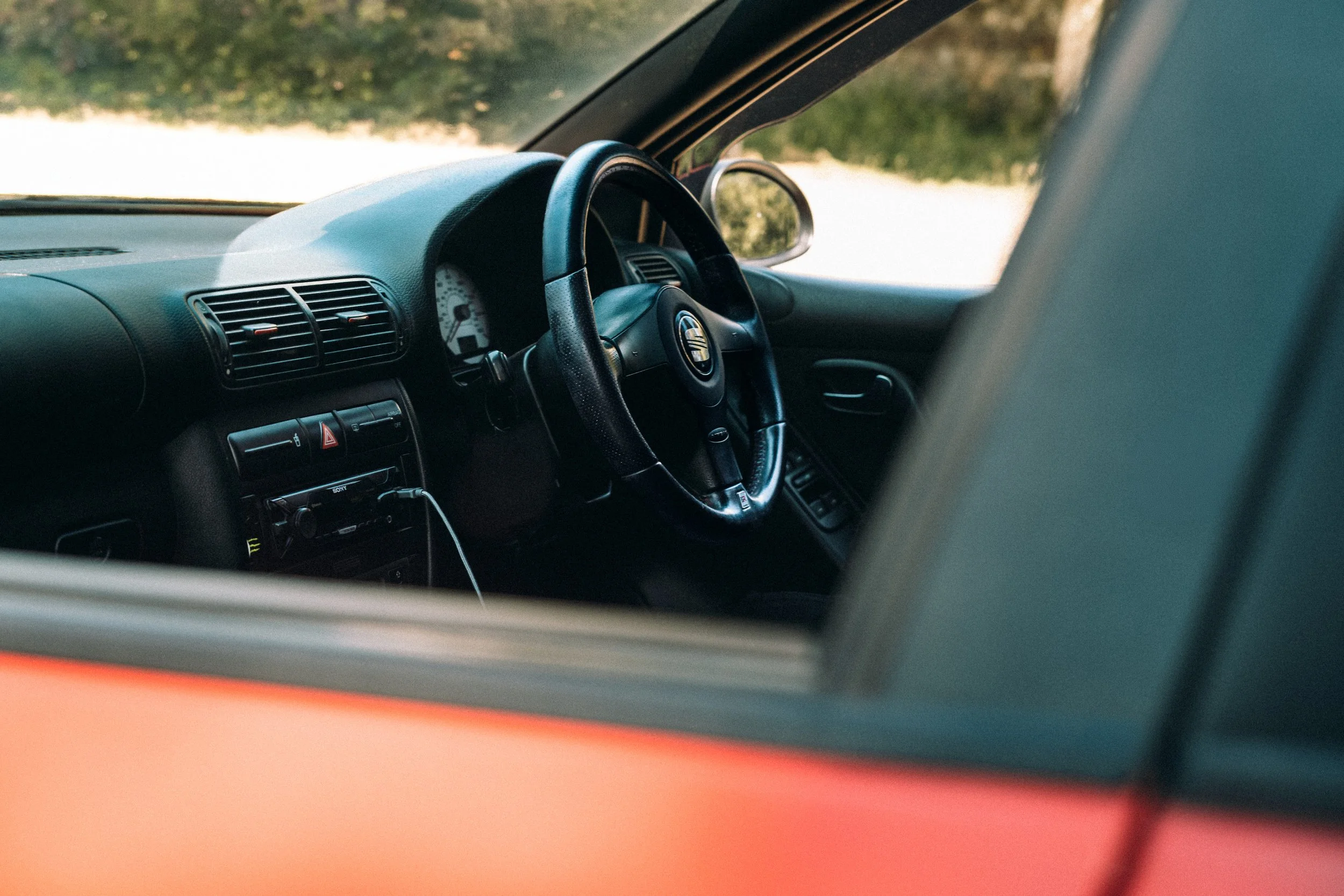 The interior of a car showing the steering wheel, dashboard, and front windshield, with an outdoor scene visible through the windshield.
