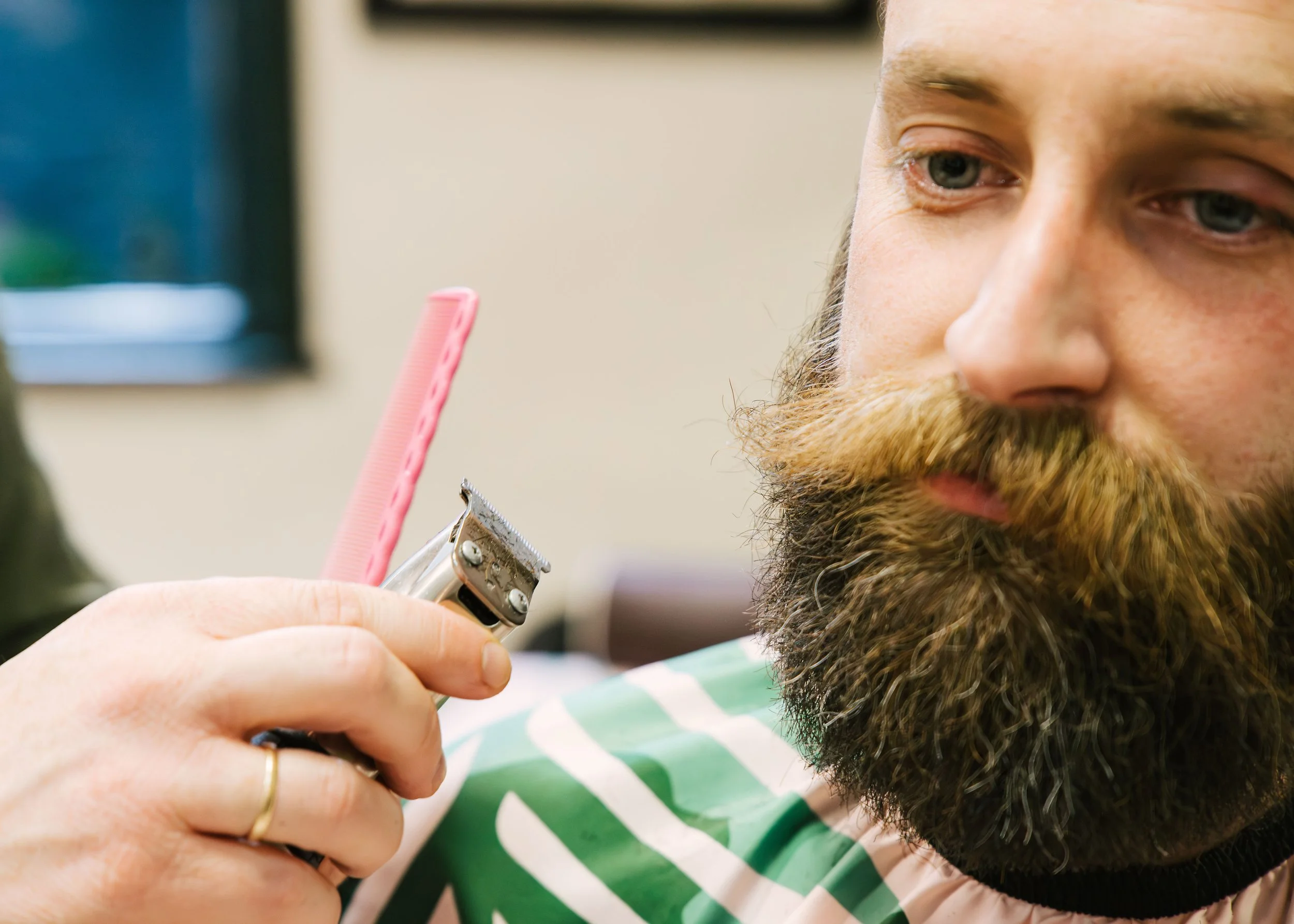 Close-up of a person with a beard receiving a haircut with electric clippers, holding a pink comb nearby, and wearing a green and white striped shirt.