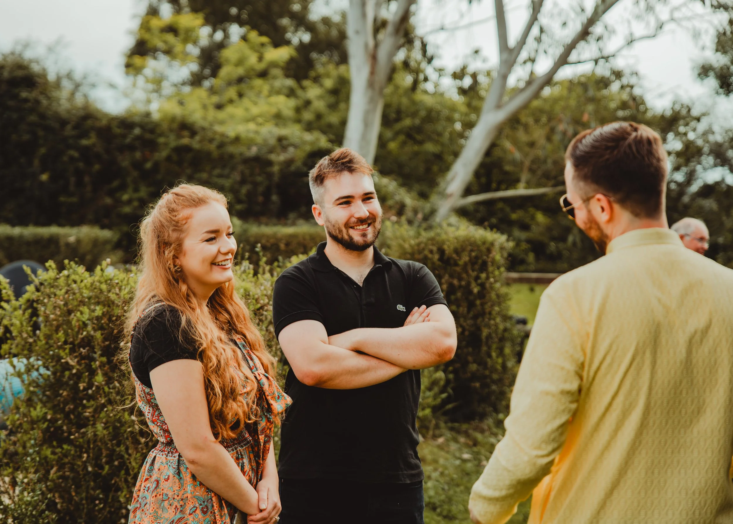 A group of four people outdoors, smiling and talking, with trees and bushes in the background.