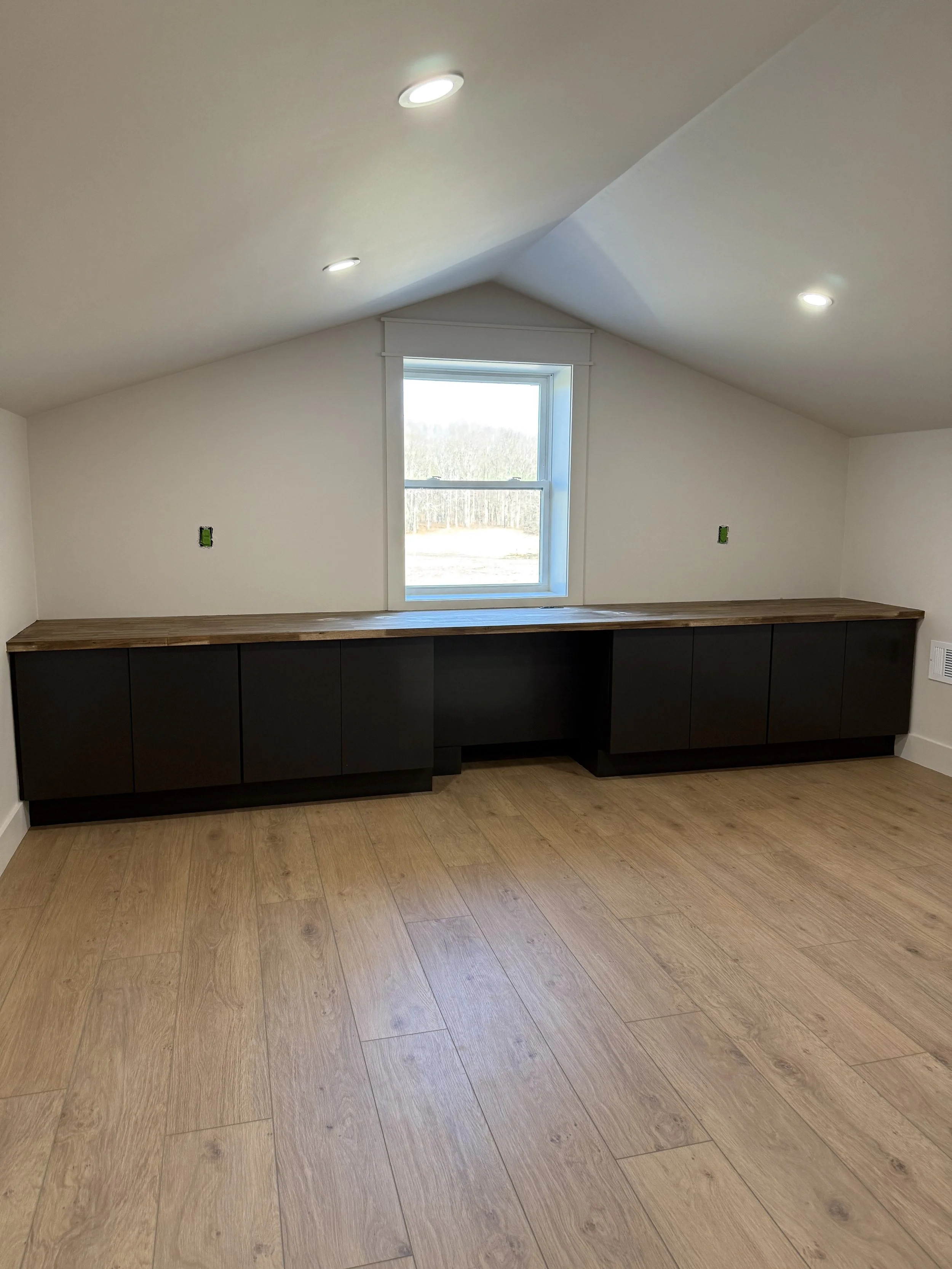 Empty room with a wooden floor, built-in black cabinet units beneath a window, white walls, and ceiling with recessed lighting.