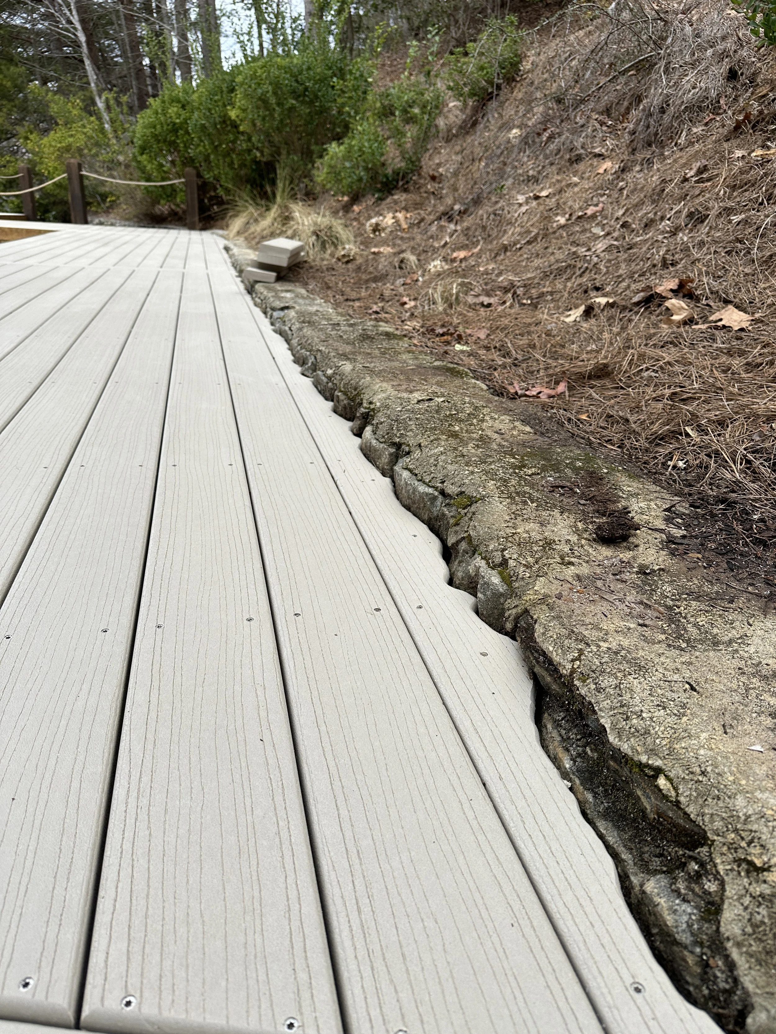 A close-up view of a wooden deck with a stone border on the side, adjacent to a soil and mulch slope with plants and trees in the background.