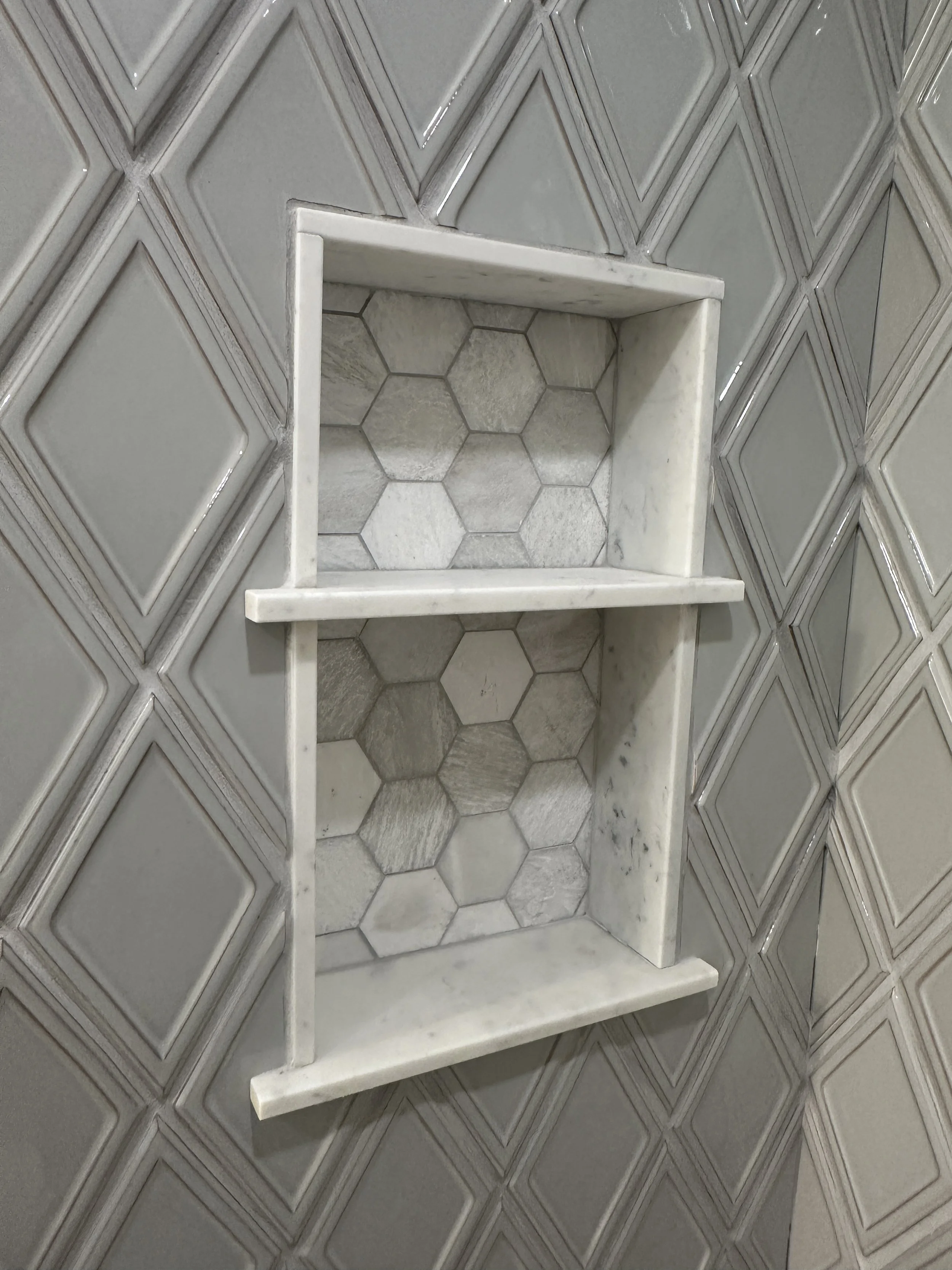 A corner of a bathroom shower wall with gray decorative tiles arranged in a diamond pattern, featuring a built-in rectangular niche with a marble frame and hexagonal marble tiles inside.