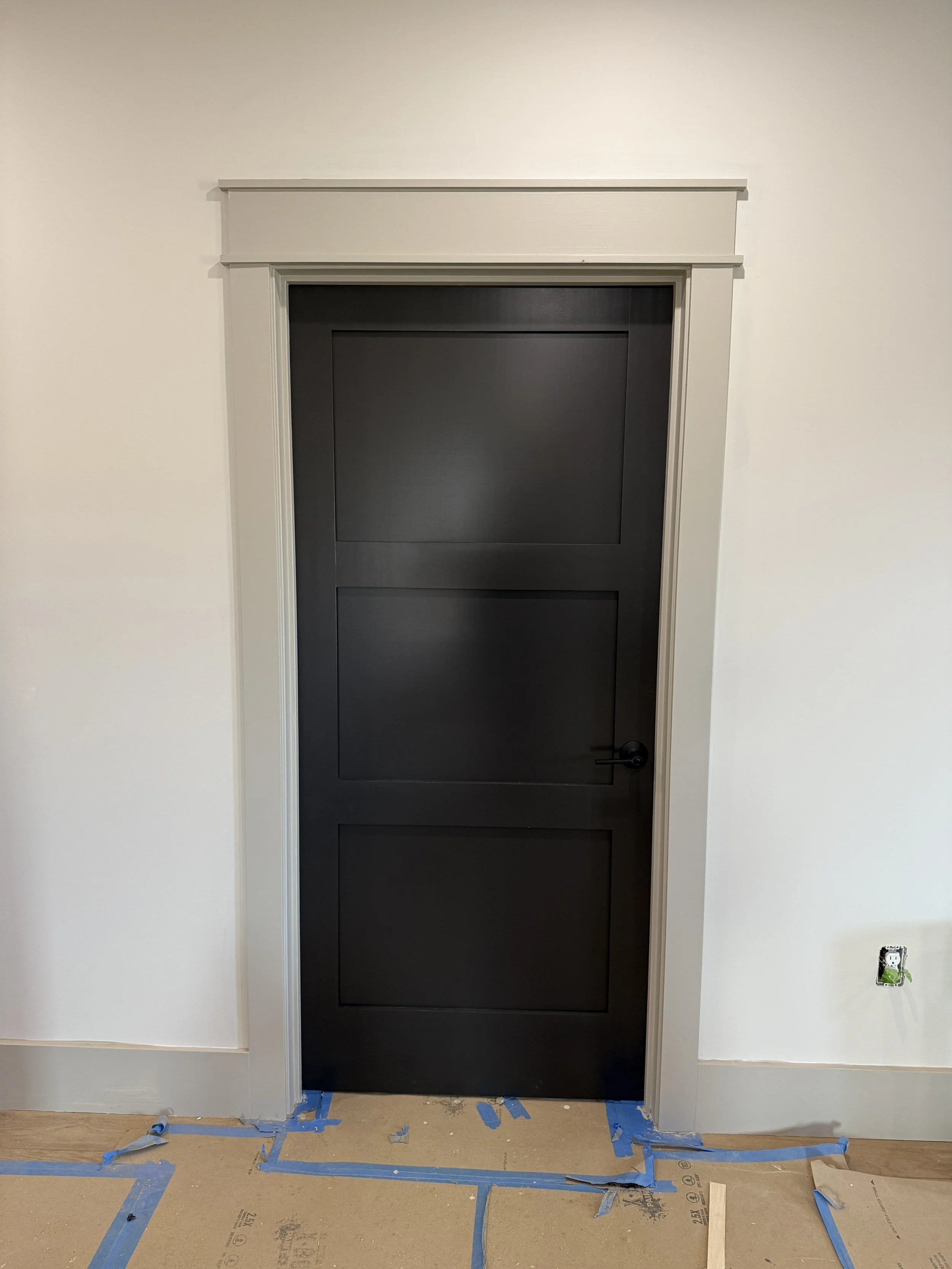 Black interior door with three rectangular panels, surrounded by white trim, partially taped on the floor of a room undergoing construction or renovation.