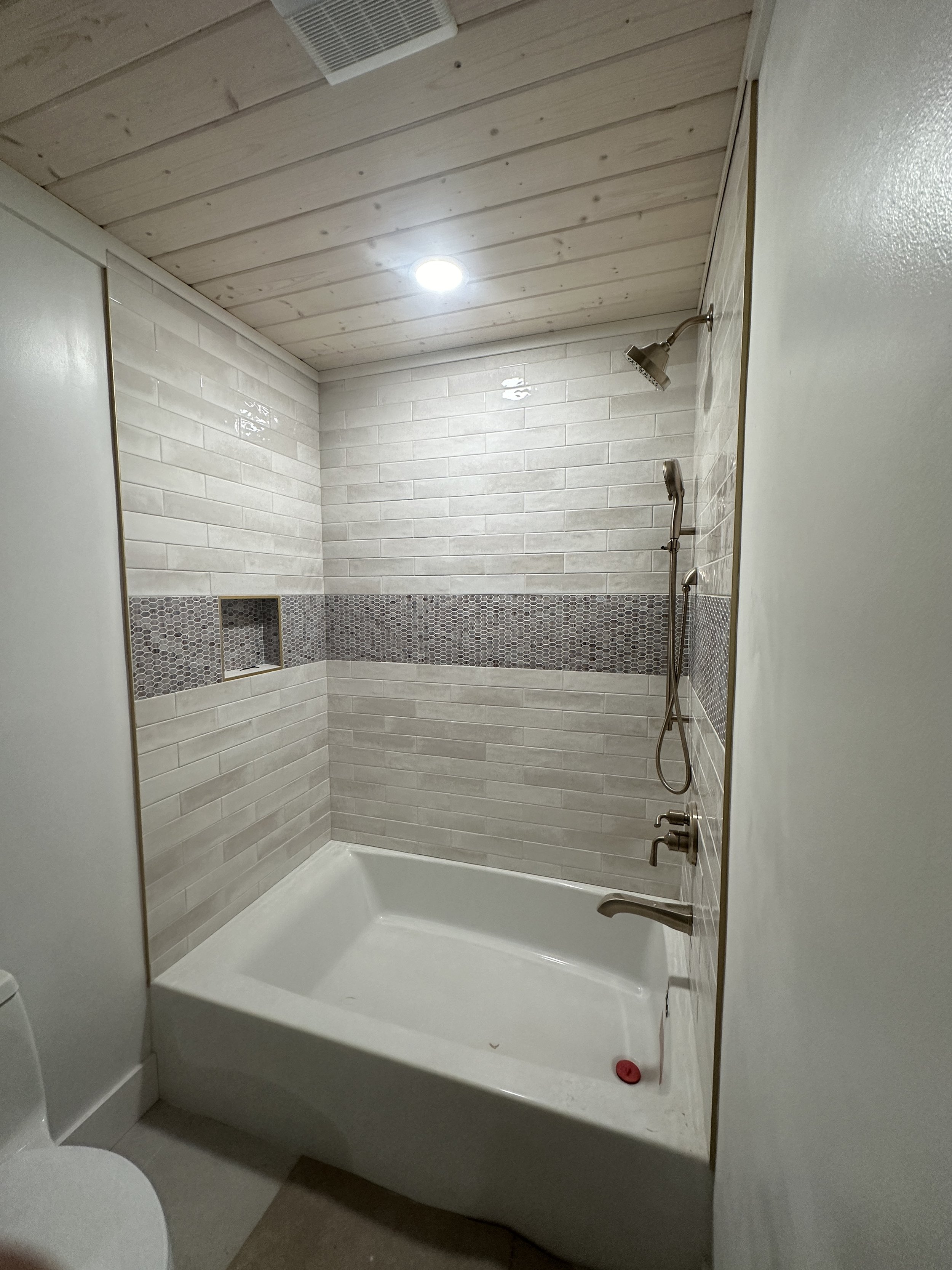 A modern bathroom with a white bathtub, beige tiled walls with a horizontal strip of small dark tiles, a wooden ceiling, and a showerhead with a handheld attachment.