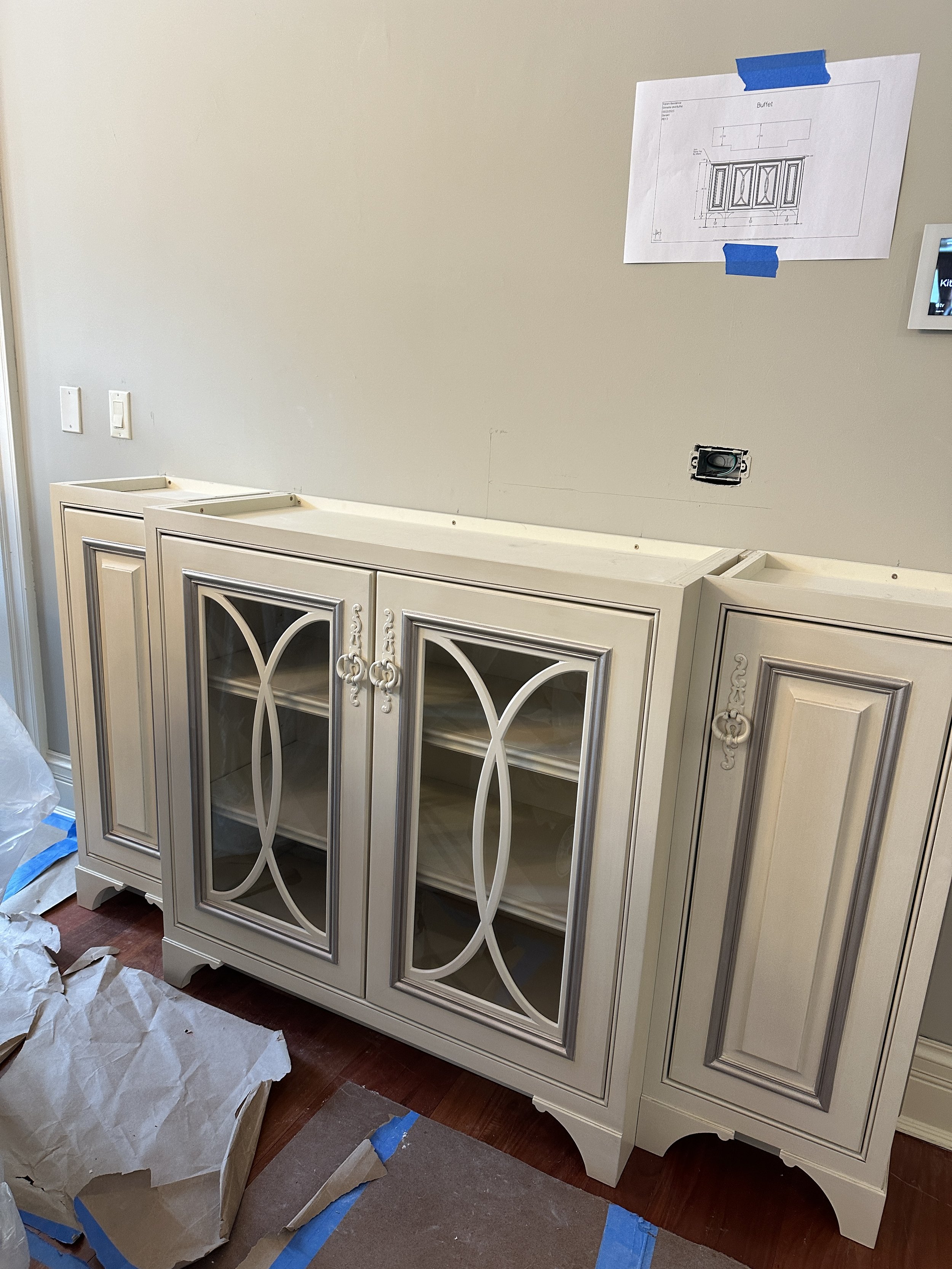 A cream-colored wooden cabinet with glass doors and decorative moldings, partially assembled in a room with construction materials on the floor and a wall with a blueprint and electrical outlets.