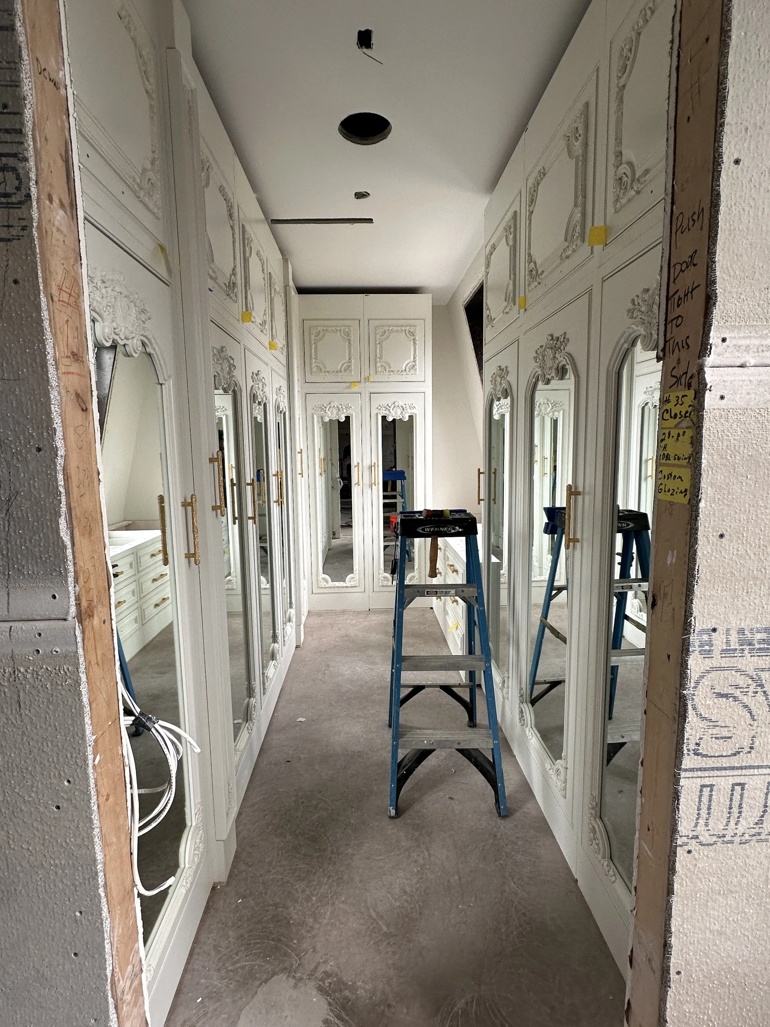 A hallway under construction with ornate white wall paneling and mirrors, a ladder in the center, and construction notes on the sides.