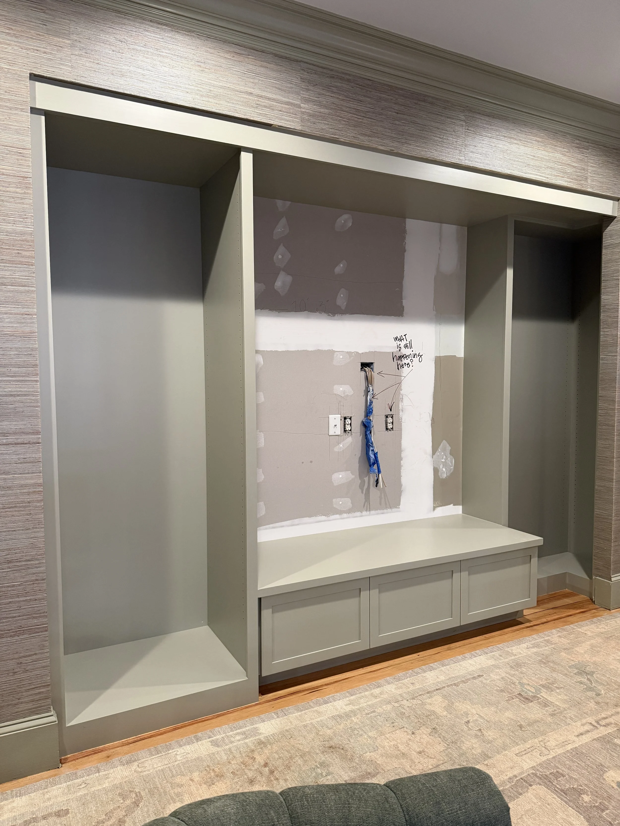 Empty built-in wall unit with cabinets, open shelving, and space for a television, in a room with carpeted floor and wall with ongoing drywall work.