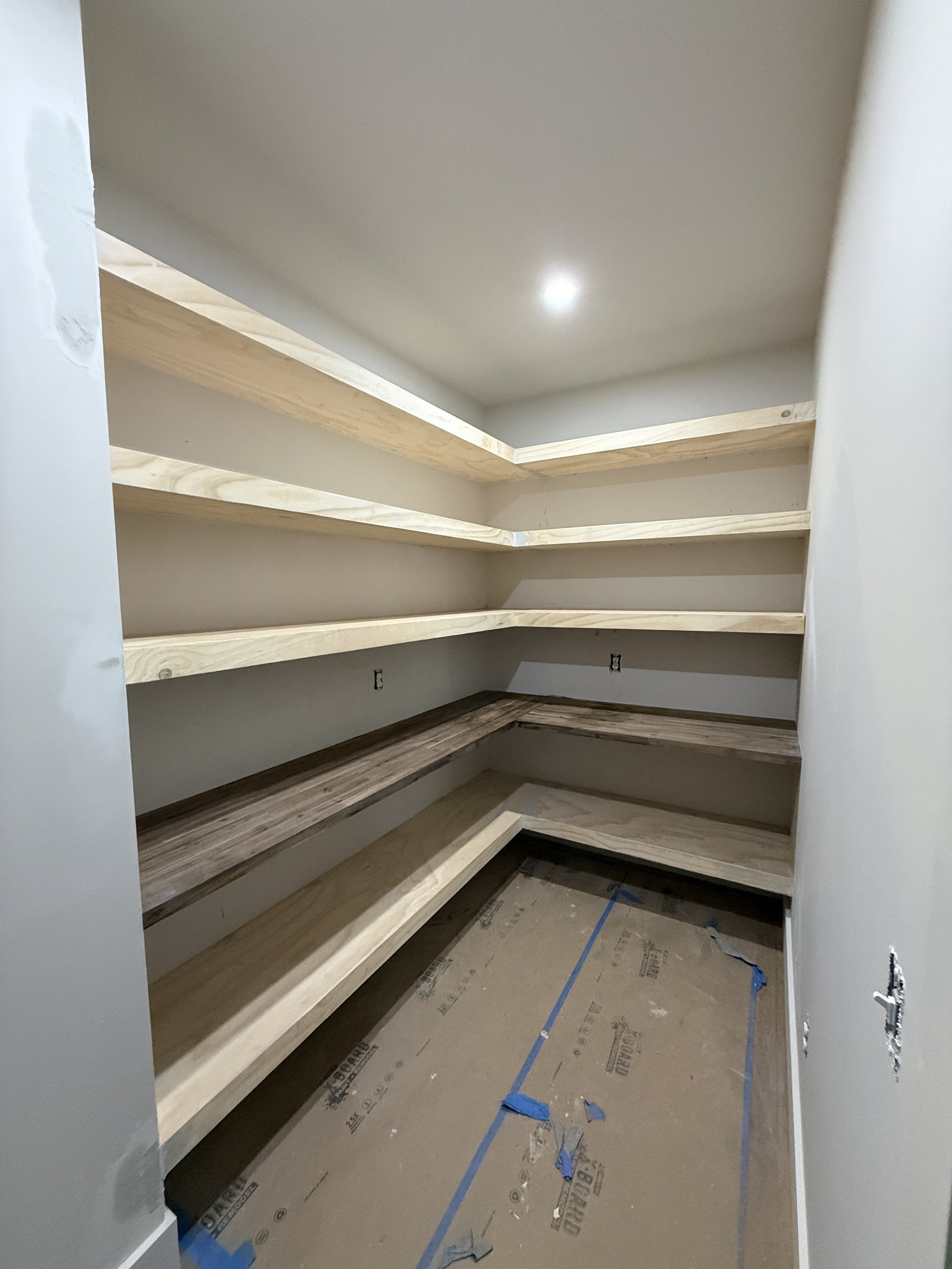 Small pantry with unfinished wooden shelves and a brown wooden countertop, with a covered concrete floor and wall outlets.