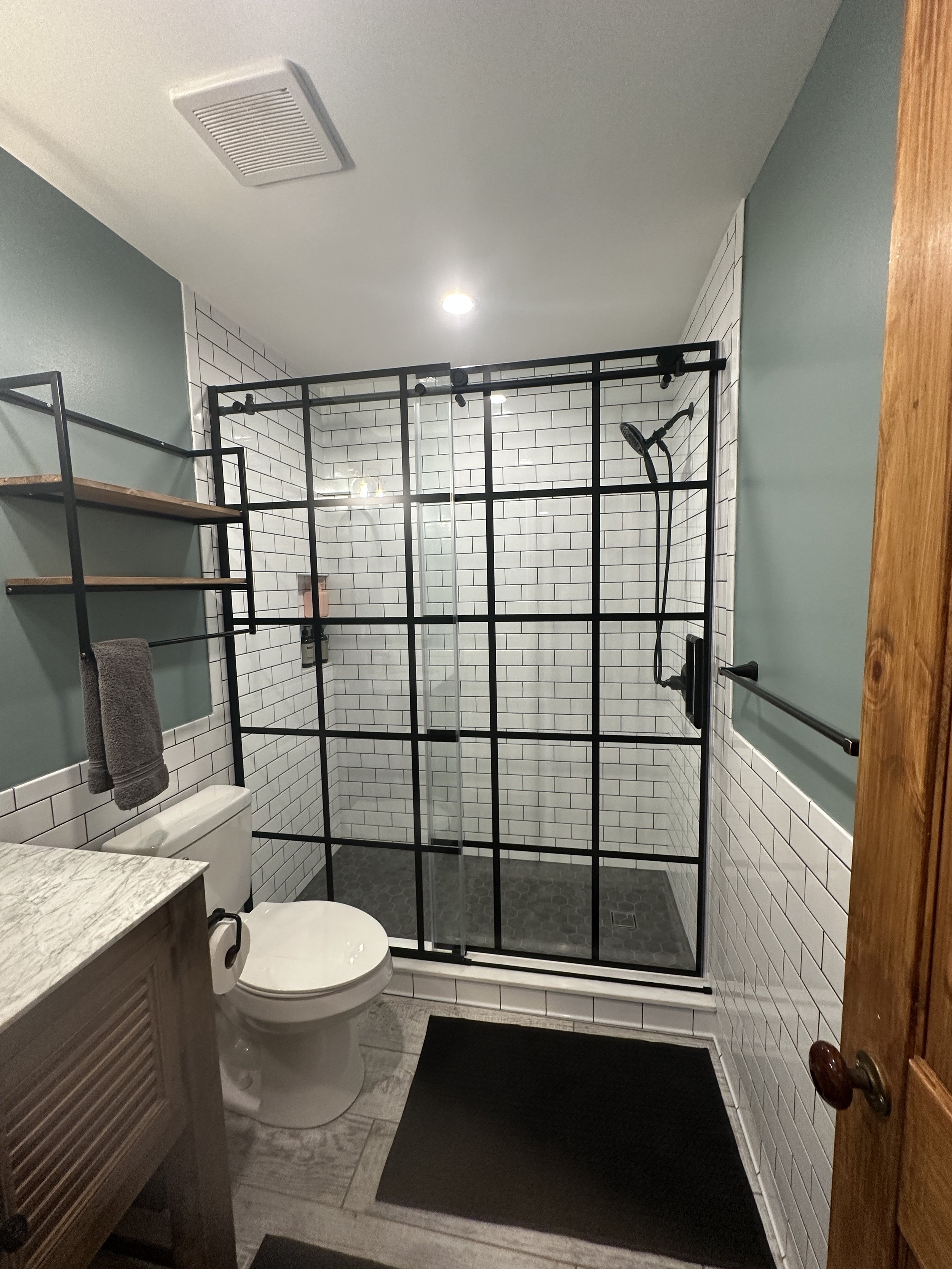 Bathroom with a glass-enclosed shower featuring black metal framing, white subway tile walls, a gray tiled shower floor, a toilet, a wooden vanity with a marble countertop, a gray towel, and shelves, with a black rug on the floor.