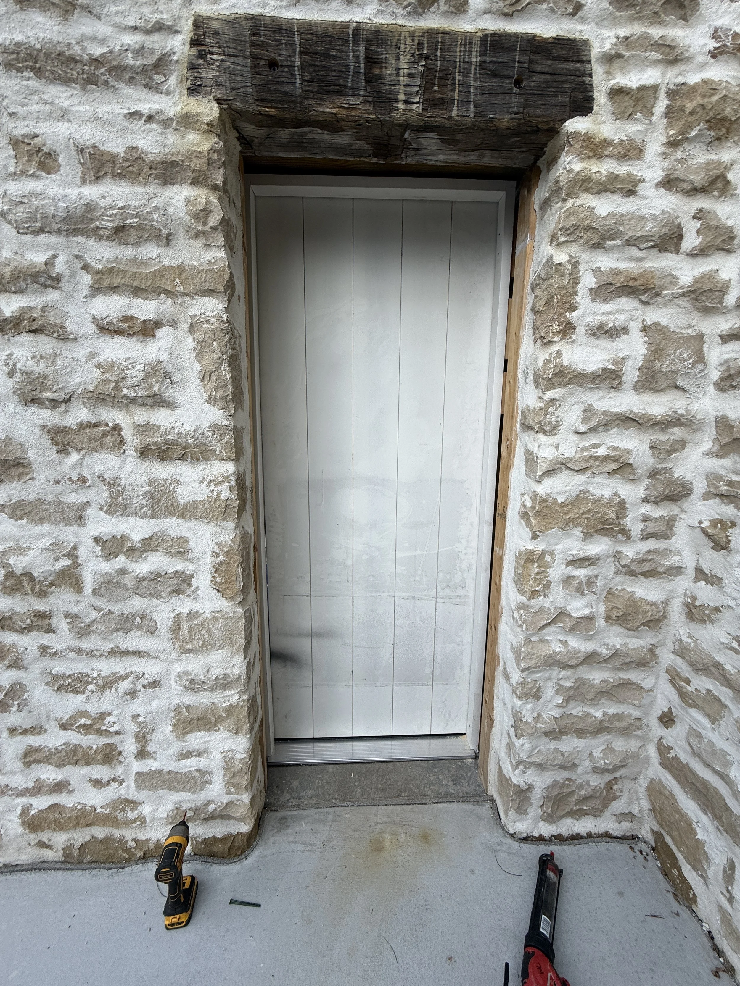A white door set in a stone wall under a rough wooden lintel, with construction tools on the ground, indicating ongoing work.