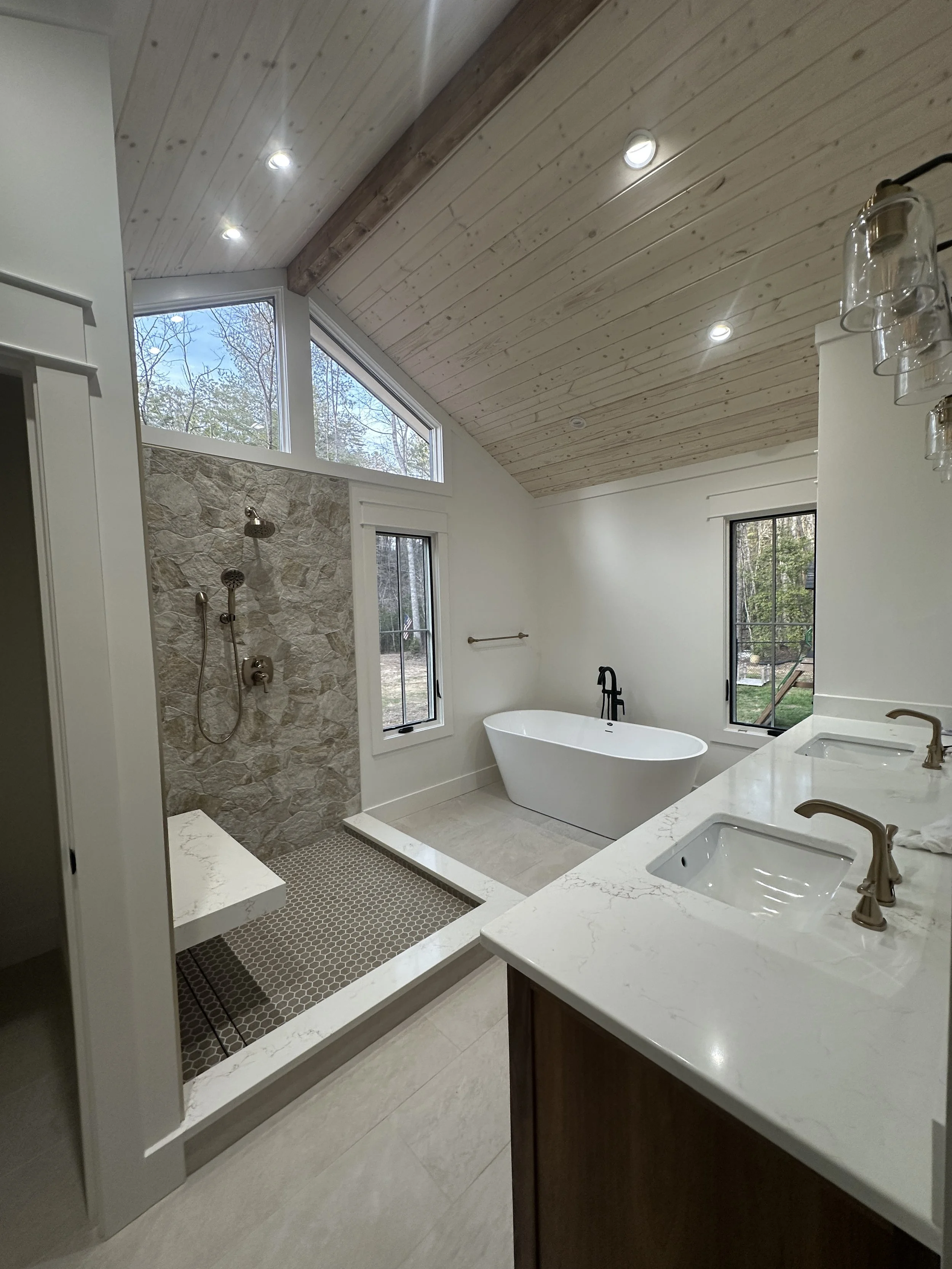 Modern bathroom with stone shower area, white bathtub near windows, dual sinks with brass fixtures, and wood-paneled ceiling with recessed lighting.