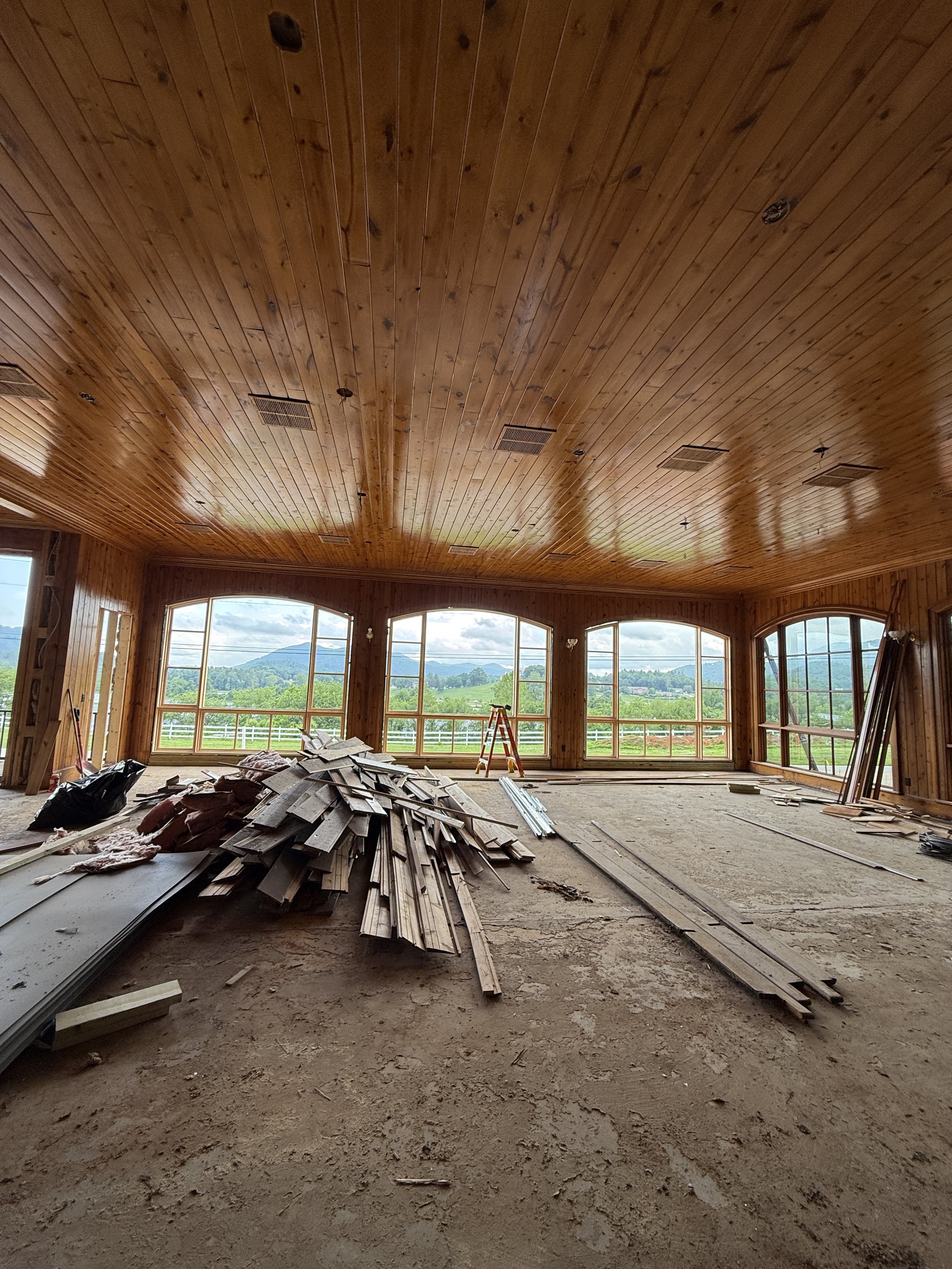 Interior of a building under construction with large windows, a wooden ceiling, and construction materials and debris on the unfinished floor.