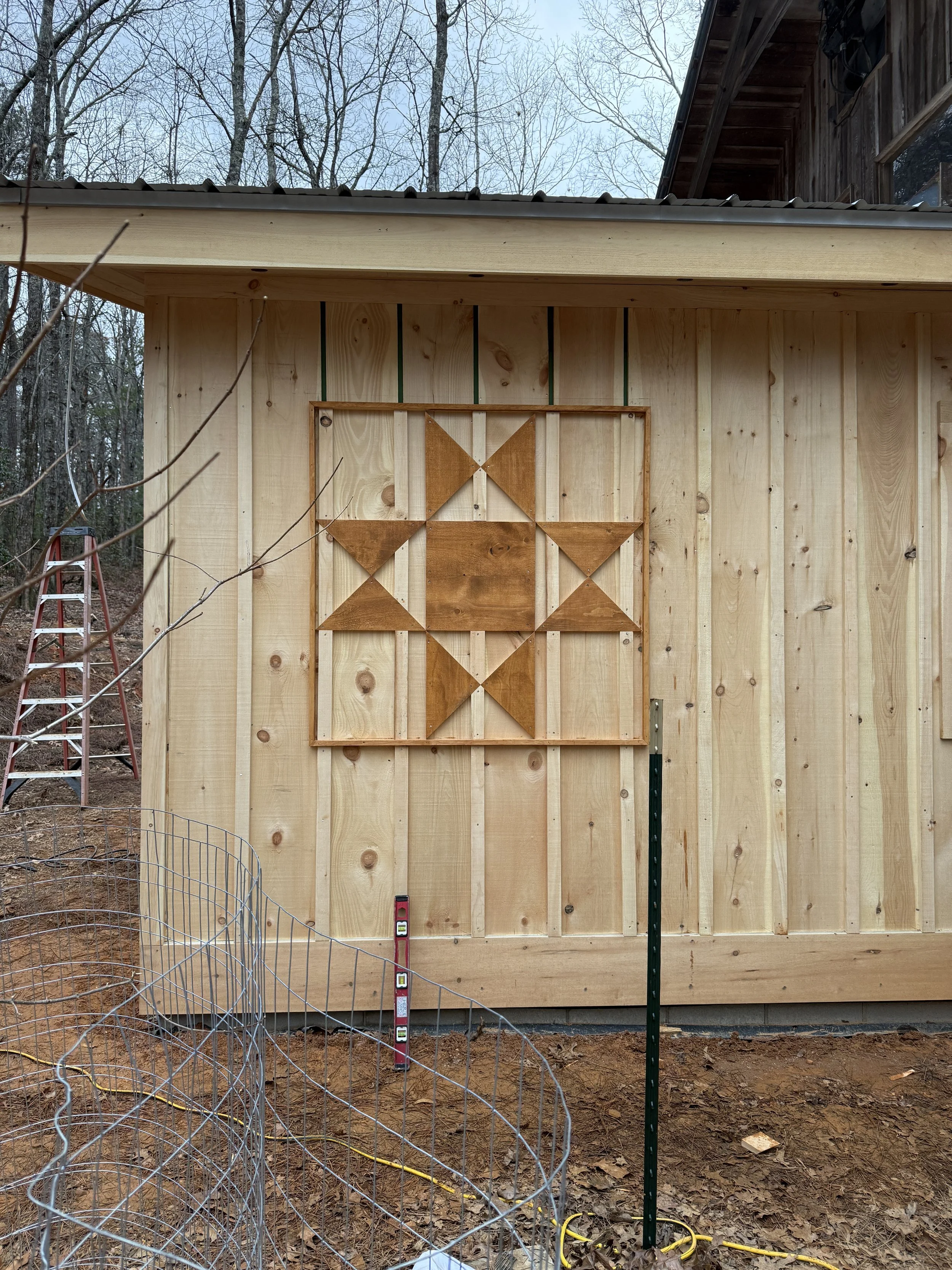 Wooden wall with decorative geometric art piece, a ladder, and construction tools in an outdoor setting with trees.