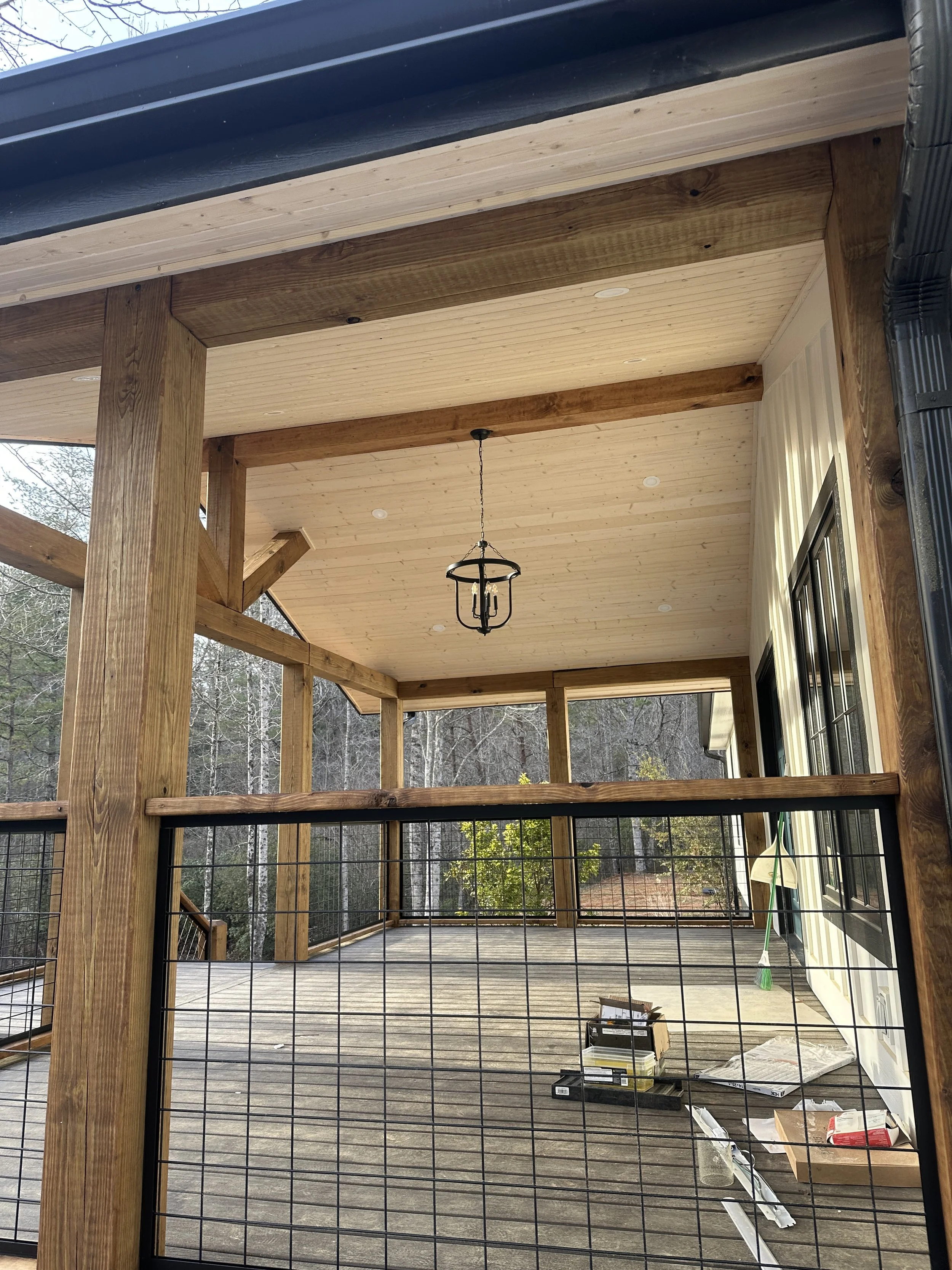 Newly built wooden porch or deck with ceiling, chandelier, and railing, with tools and materials on the floor and a broom leaning against the house wall.
