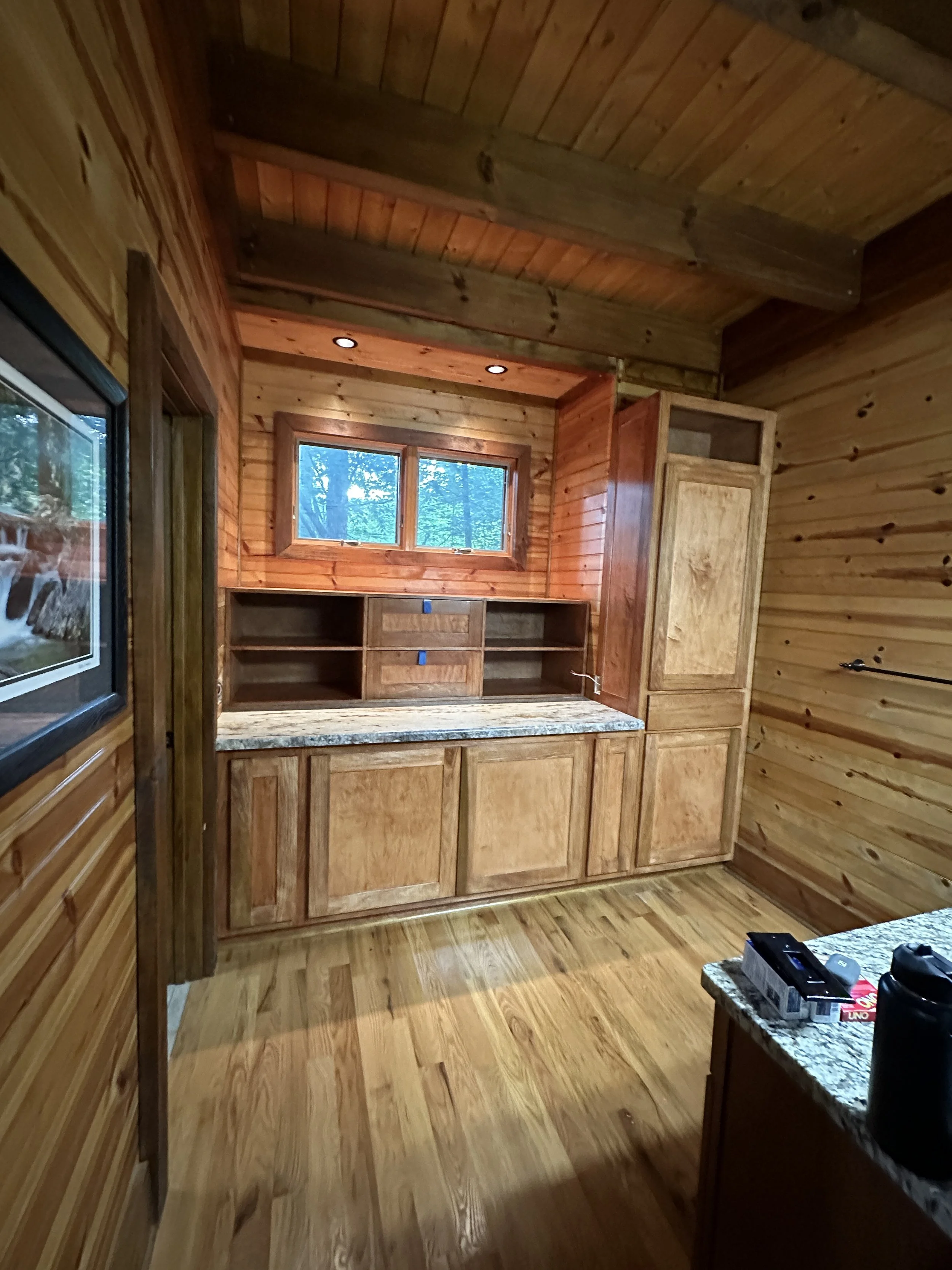 Interior view of a wooden kitchen with natural wood cabinets, granite countertop, and a small window above the workspace, with tools and supplies on the counter.