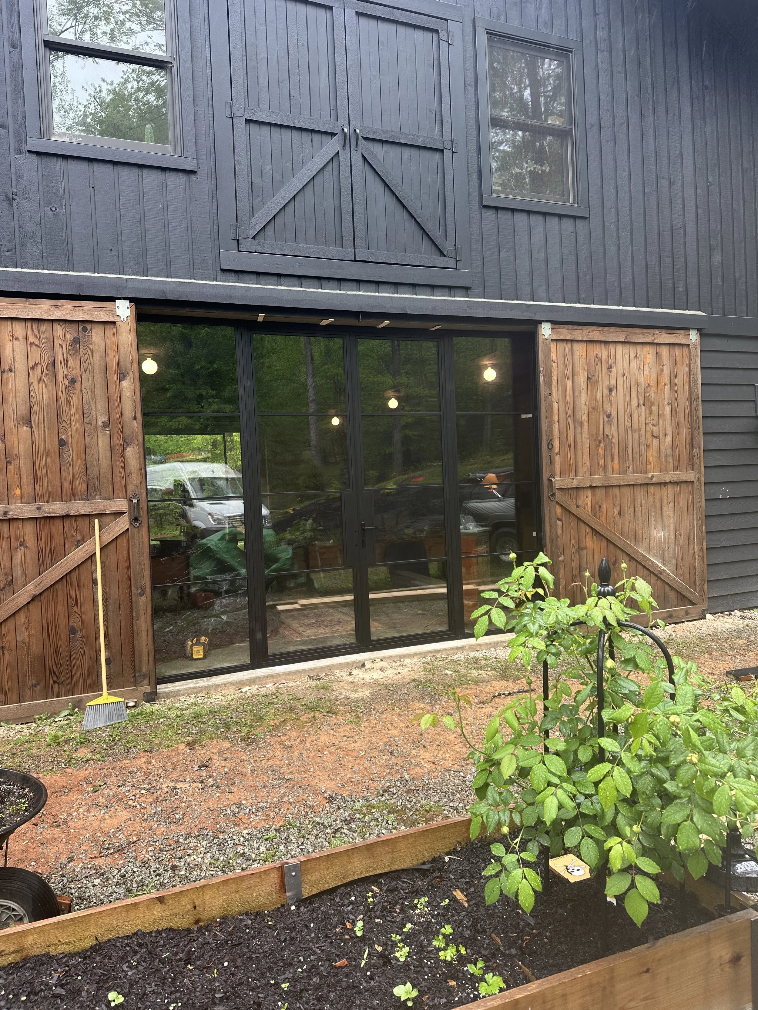A black wooden house with two glass doors and brown barn-style sliding doors. There are trees, cars, and lights reflected in the glass doors, and a small garden bed with a green leafy plant and mulch in the foreground.