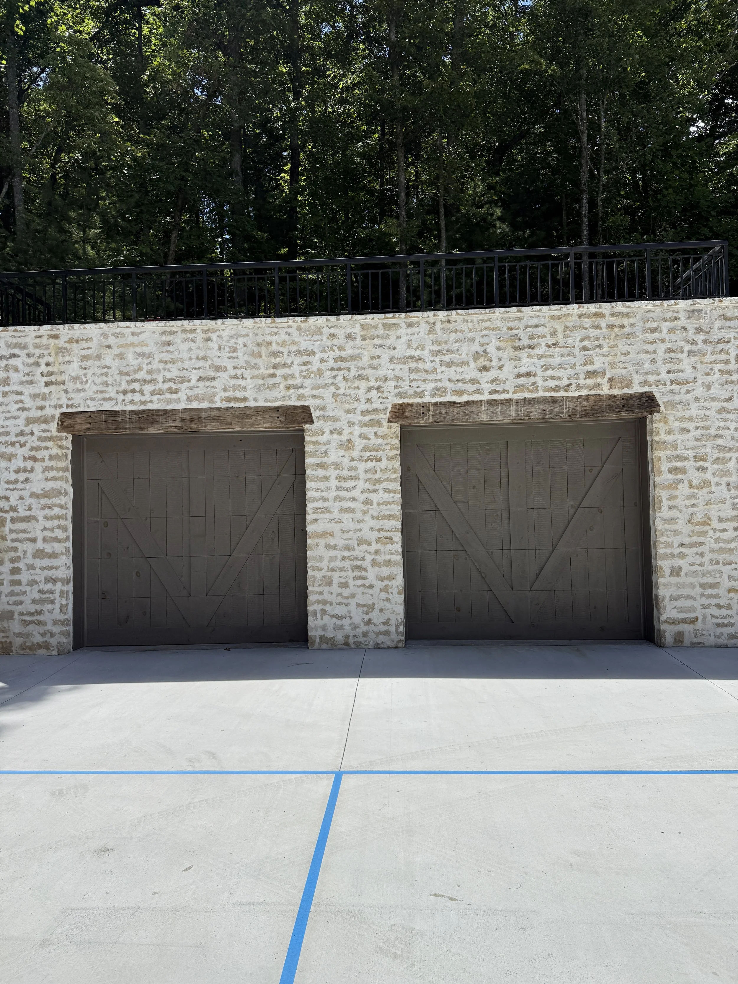 Two dark wooden garage doors set into a stone wall with a concrete driveway in front, surrounded by tall trees and greenery.