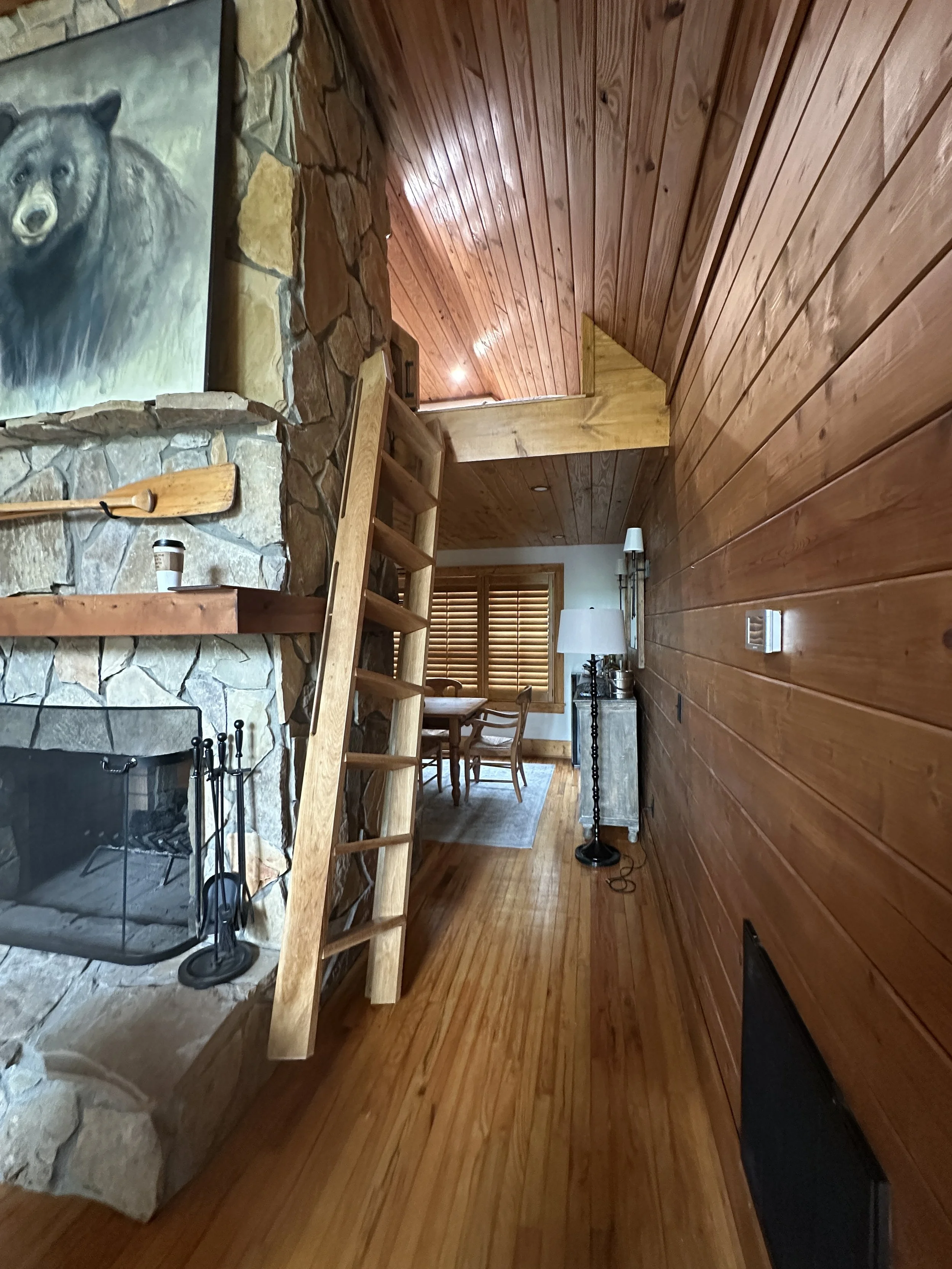Interior view of a cozy cabin with wooden walls and floor, a stone fireplace, a wooden ladder leading to a loft, a dining table with chairs, and a standing lamp near a window with wooden blinds.