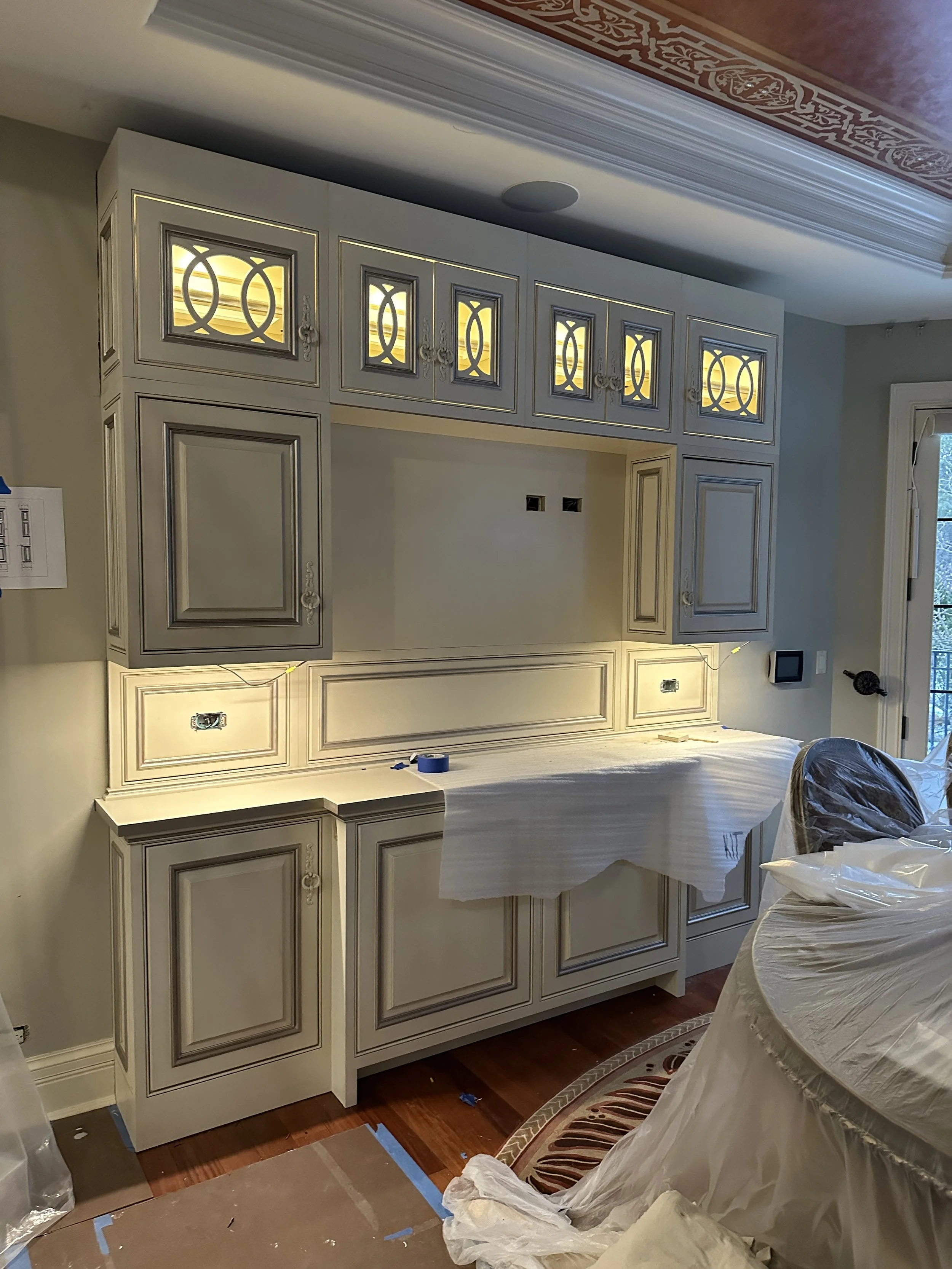 Kitchen cabinetry installation in progress, featuring cream-colored cabinets with decorative molding and frosted glass panel doors, some parts covered with plastic and masking tape, a window to the right, and construction materials on the floor.