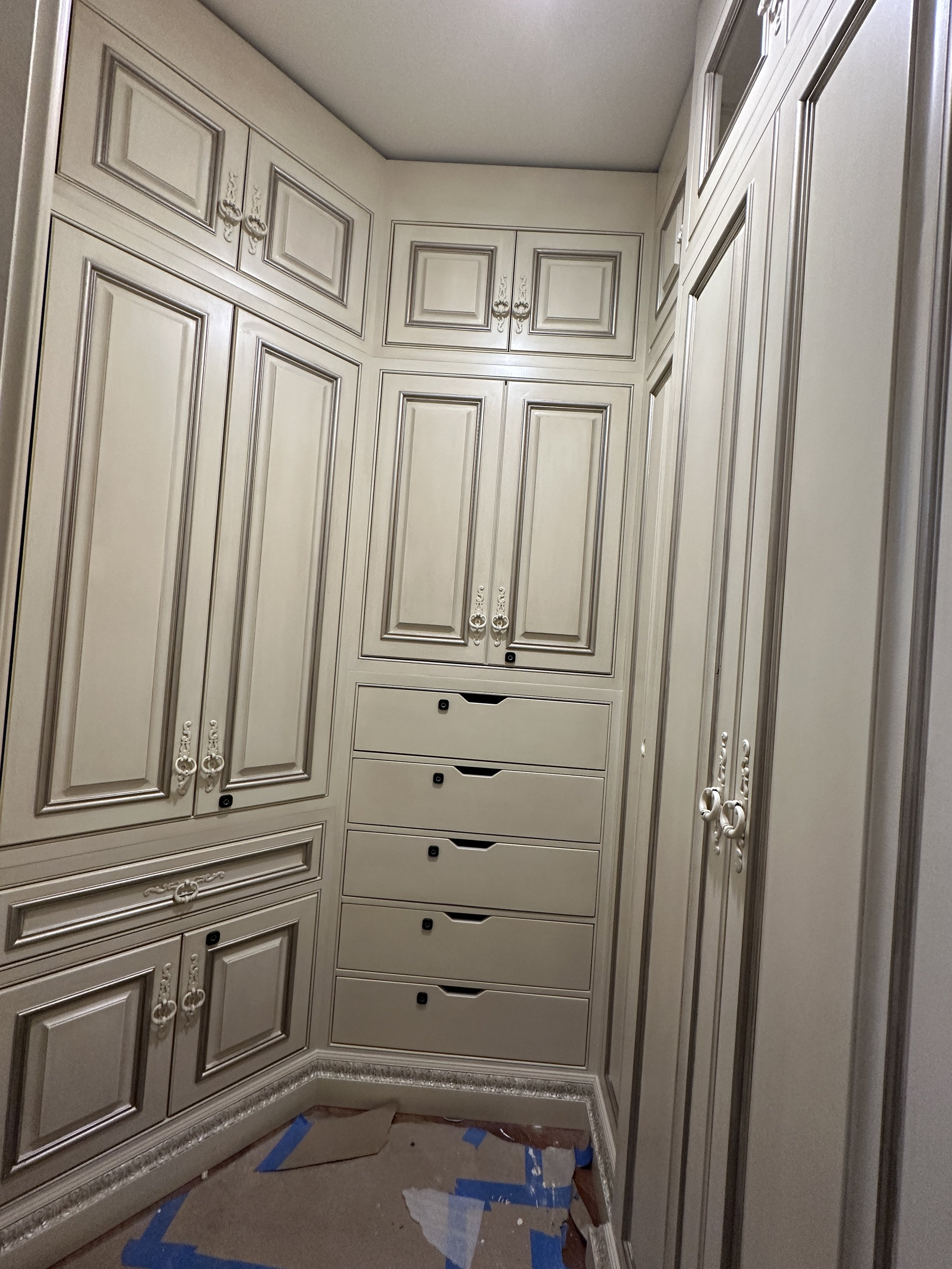 Built-in wooden cabinetry with ornate handles in a corner of a room, with drawers and cabinet doors, painted in light beige color.