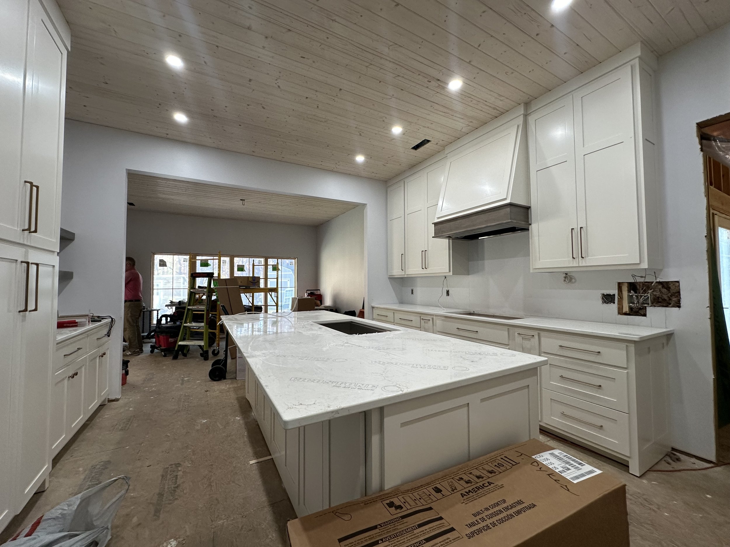 Modern kitchen under construction with white cabinets, a large marble island, and a wooden ceiling with recessed lighting. Workers and construction materials are visible in the background.