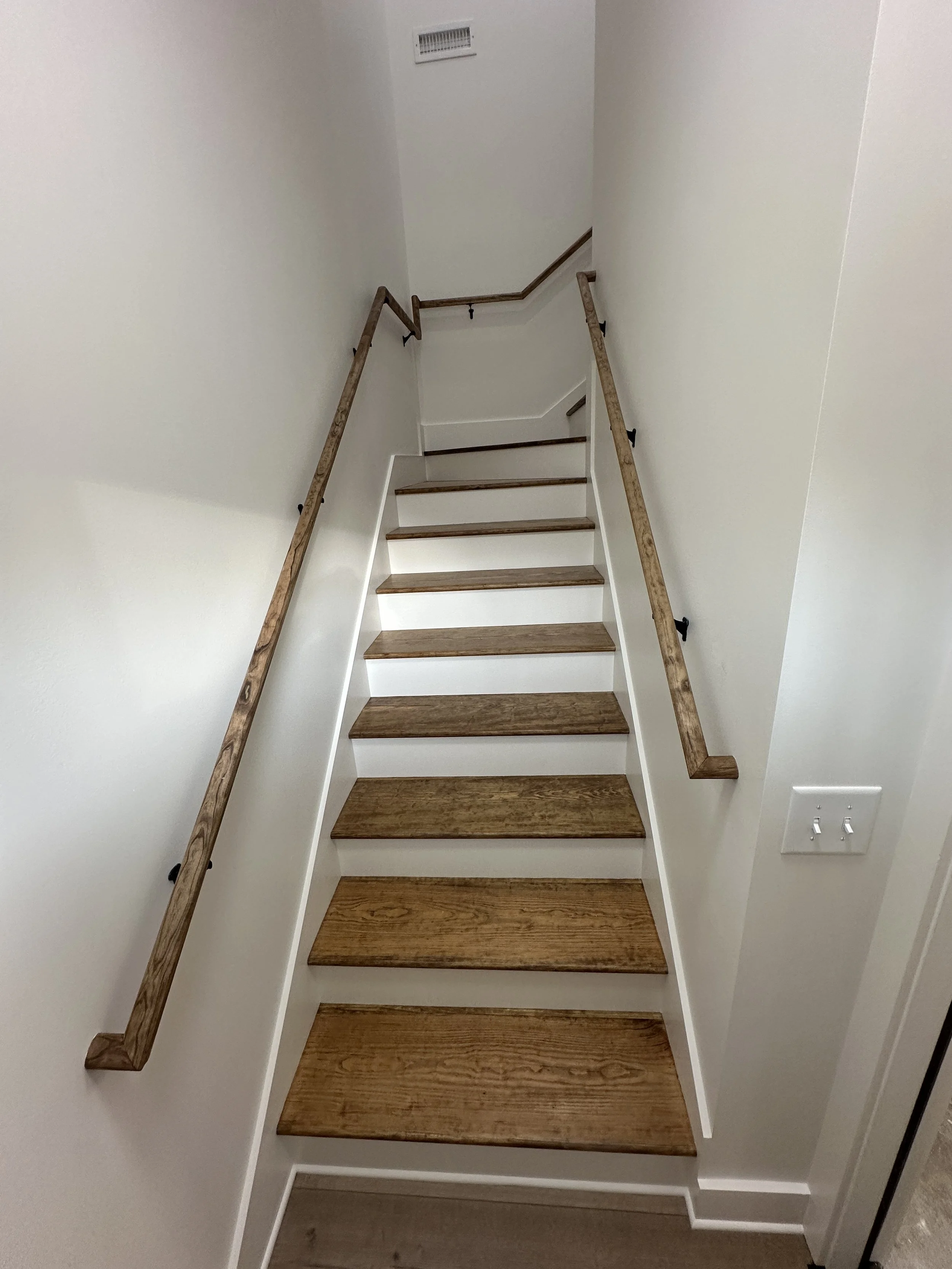Interior view of a staircase with wooden steps, white risers, and wooden handrails on both sides, leading upward.