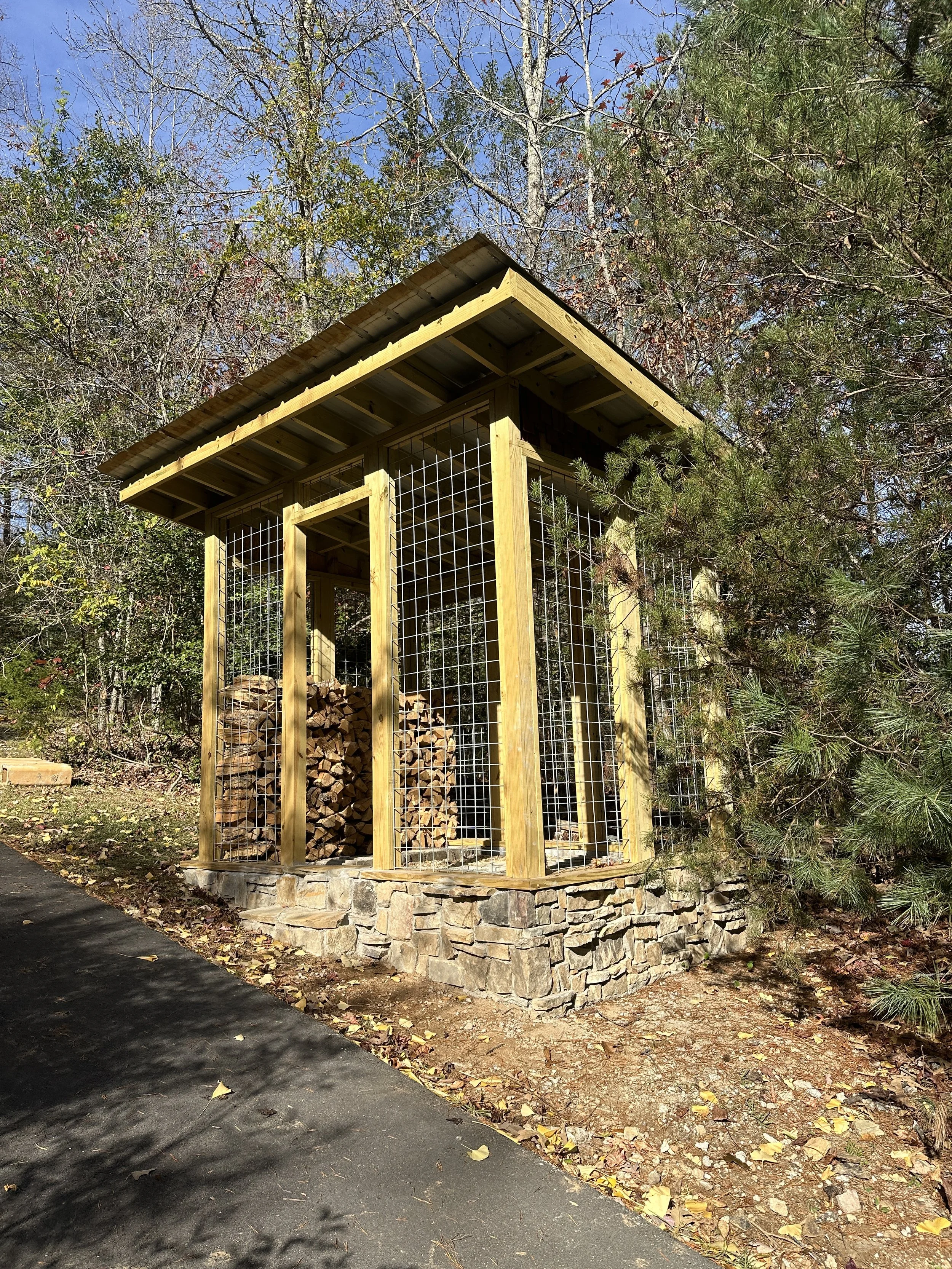 Wood storage shed with stone foundation and wire mesh walls, filled with chopped firewood, surrounded by trees and situated beside a paved path.