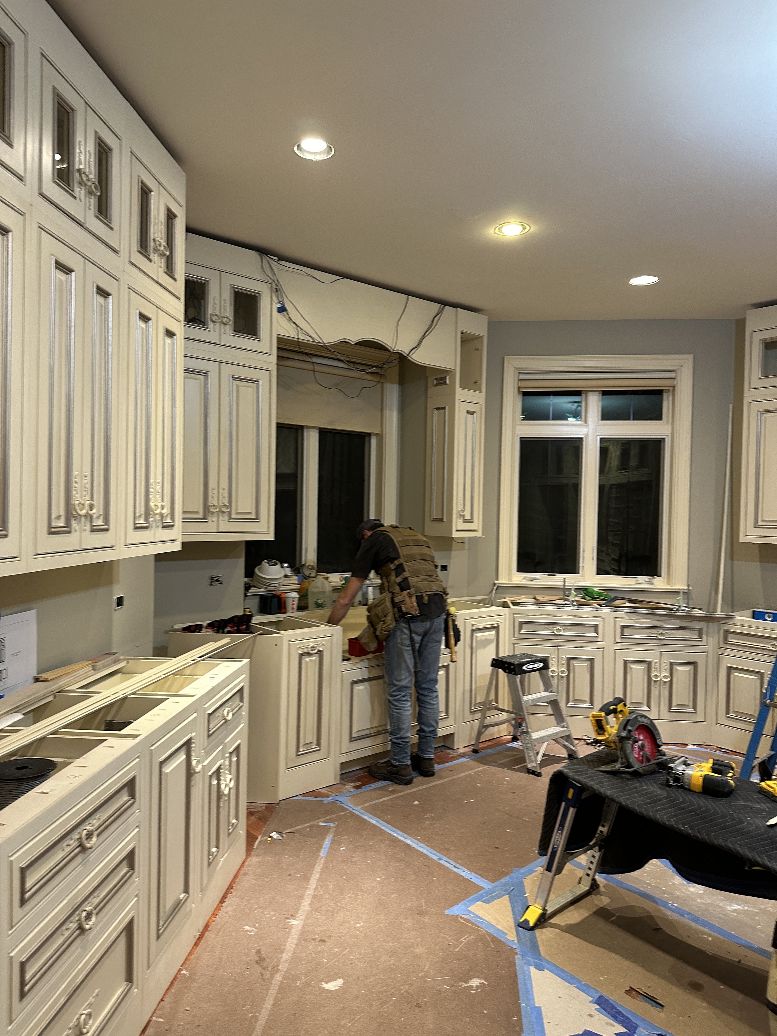 A person working in a kitchen under renovation, with beige cabinets, a window, and construction tools on the floor.