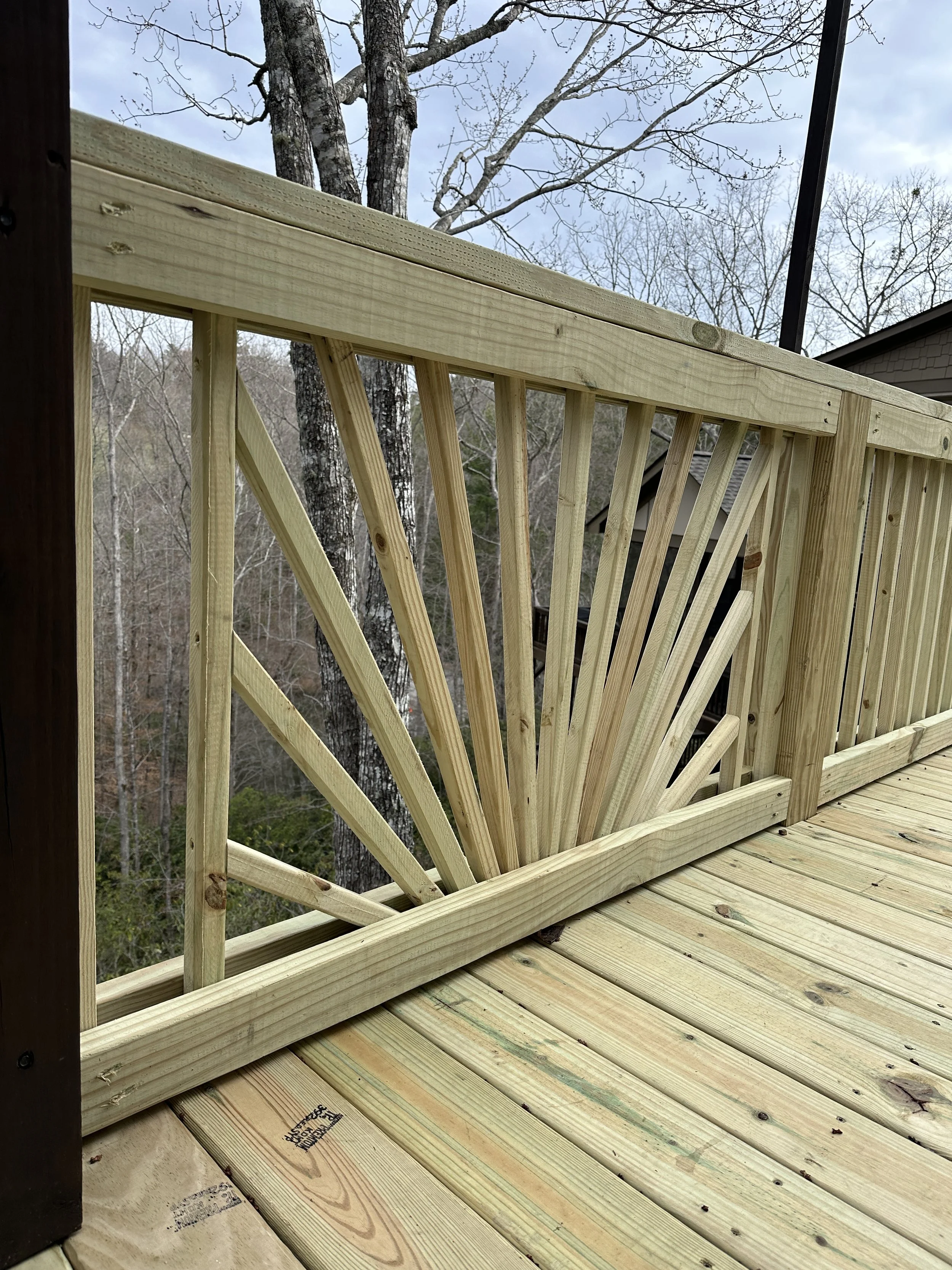Wooden deck with a decorative railing design, surrounded by trees and a cloudy sky.