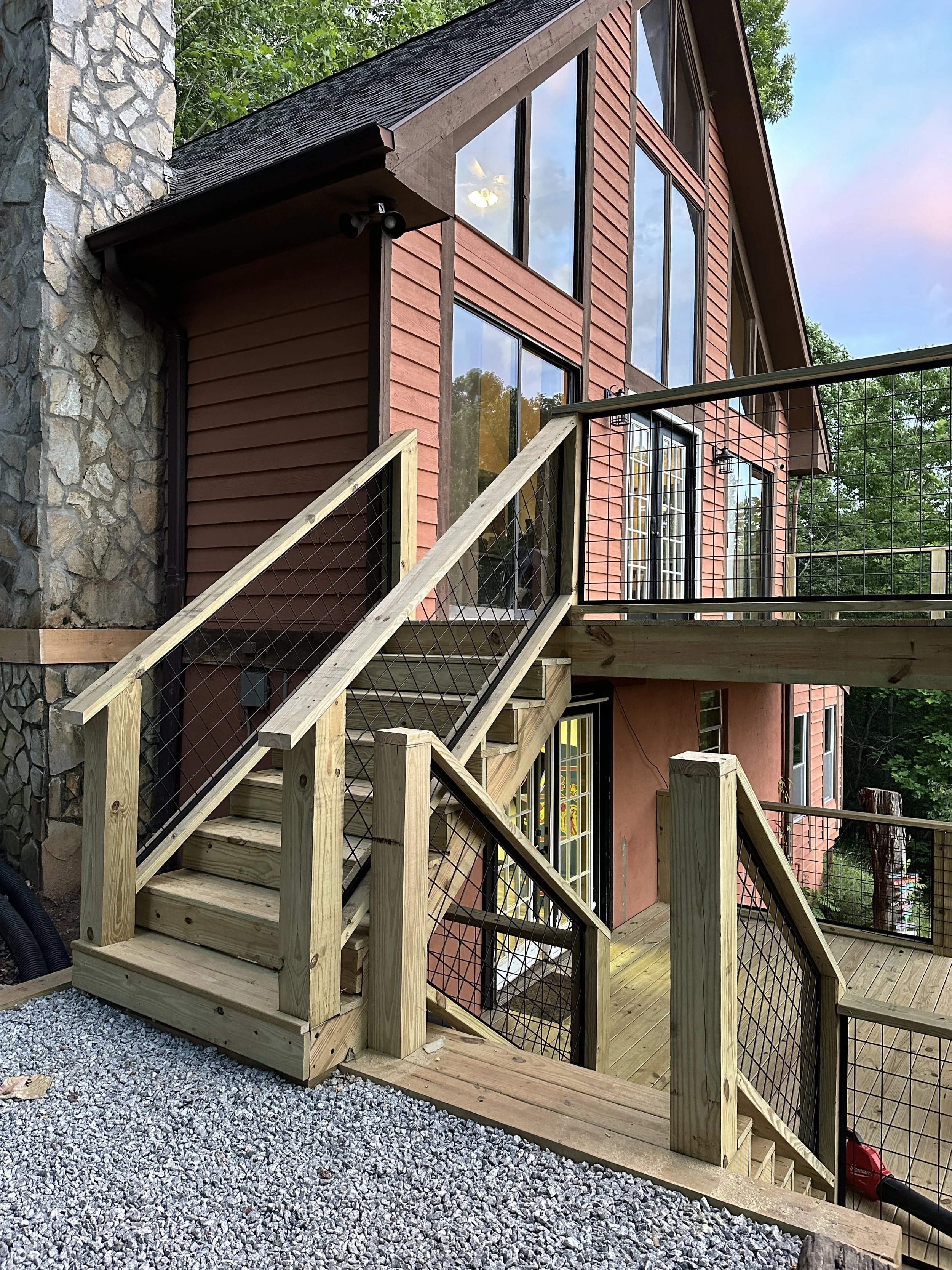 Newly constructed outdoor wooden staircase with metal railings leading to a deck outside a multi-story house with large windows, red siding, and stone chimney, surrounded by trees.