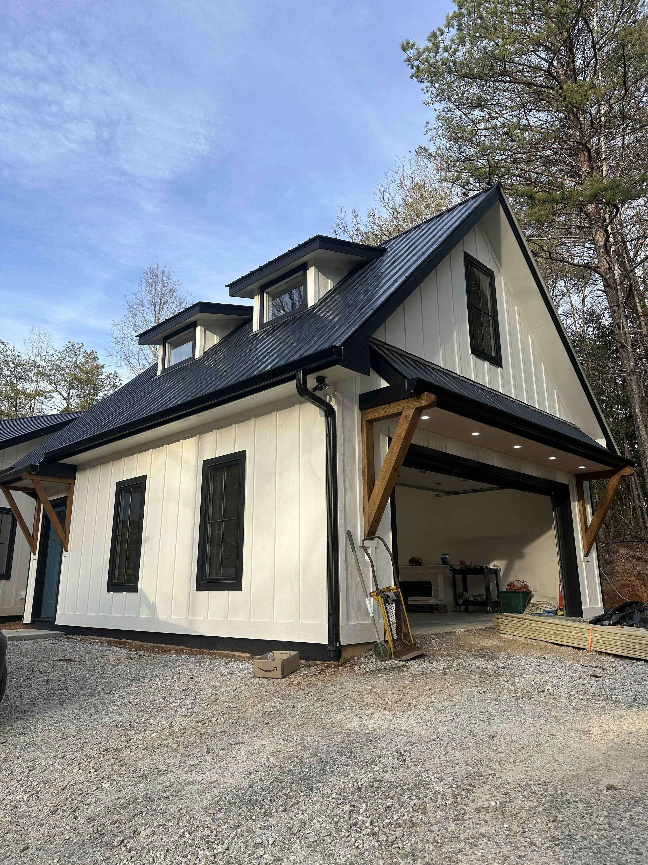 Newly constructed modern two-story house with black metal roof, white vertical siding, and black-framed windows. The garage is open, showing renovation tools inside, while wooden support beams are visible at the entrance. The house is surrounded by a