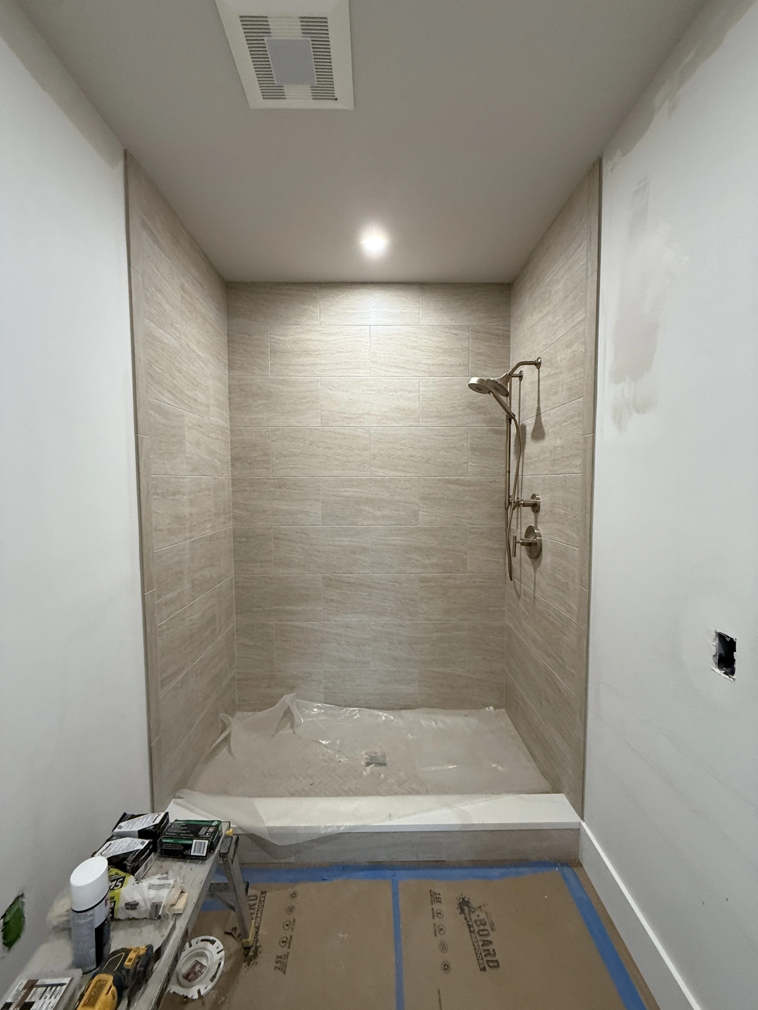 Bathroom shower area under construction with beige tiled walls, a showerhead, and construction tools and supplies on a nearby table.