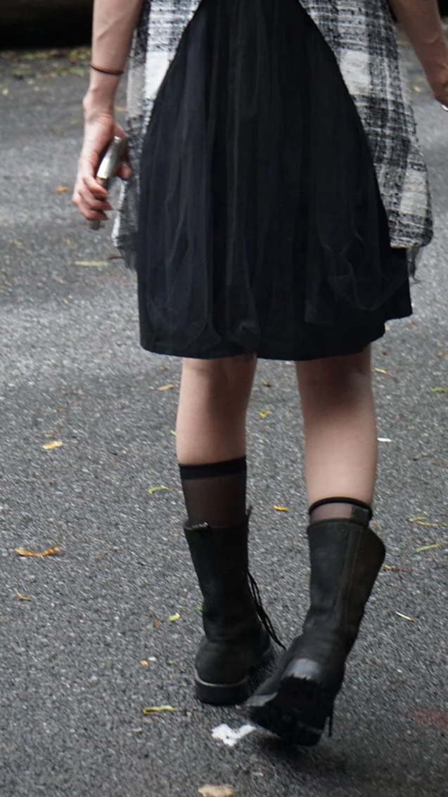 A person wearing black boots, black sheer socks, a black skirt, and a plaid shirt, standing on a paved surface with fallen leaves, holding something in their right hand.