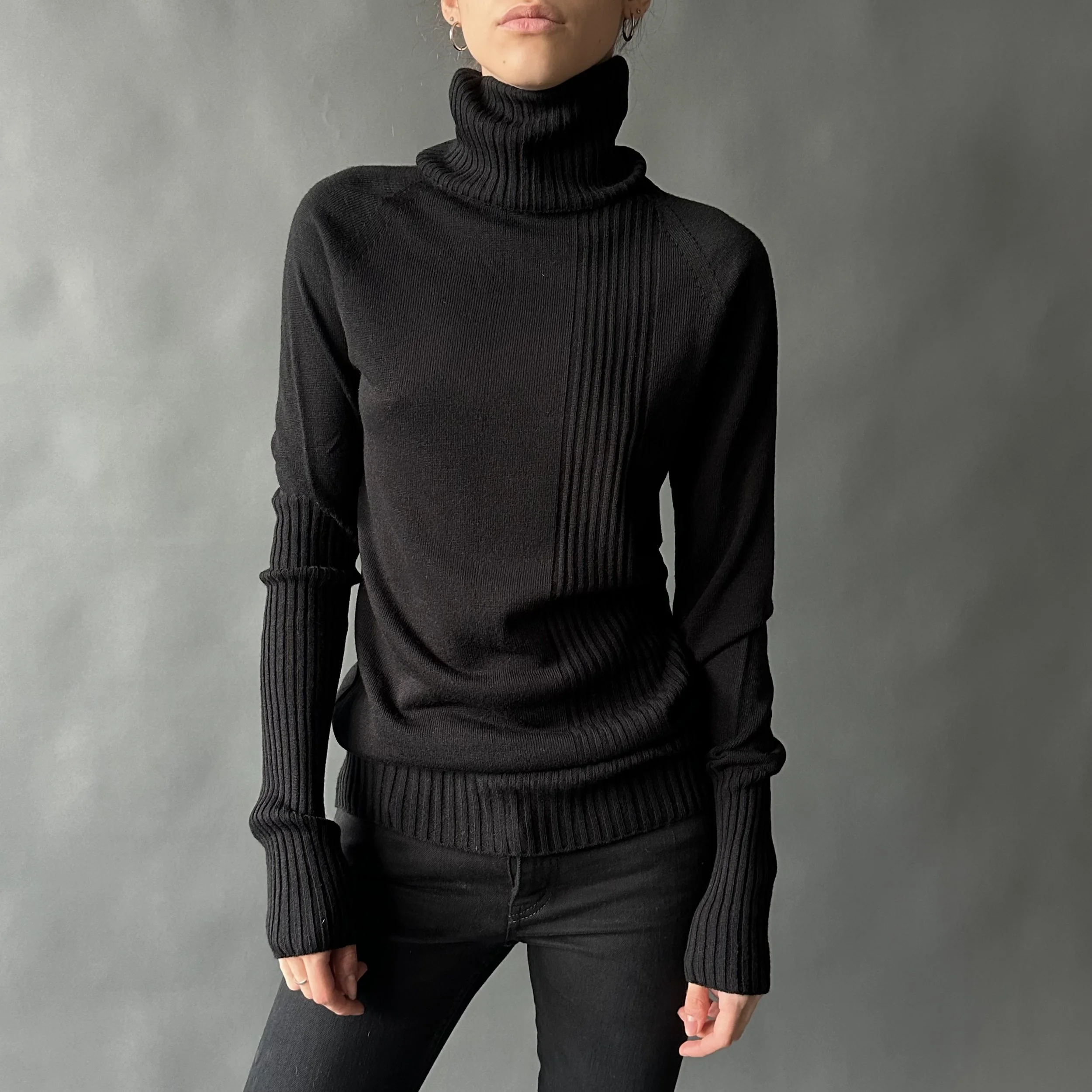 Person wearing a black turtleneck sweater and black pants standing against a gray background.