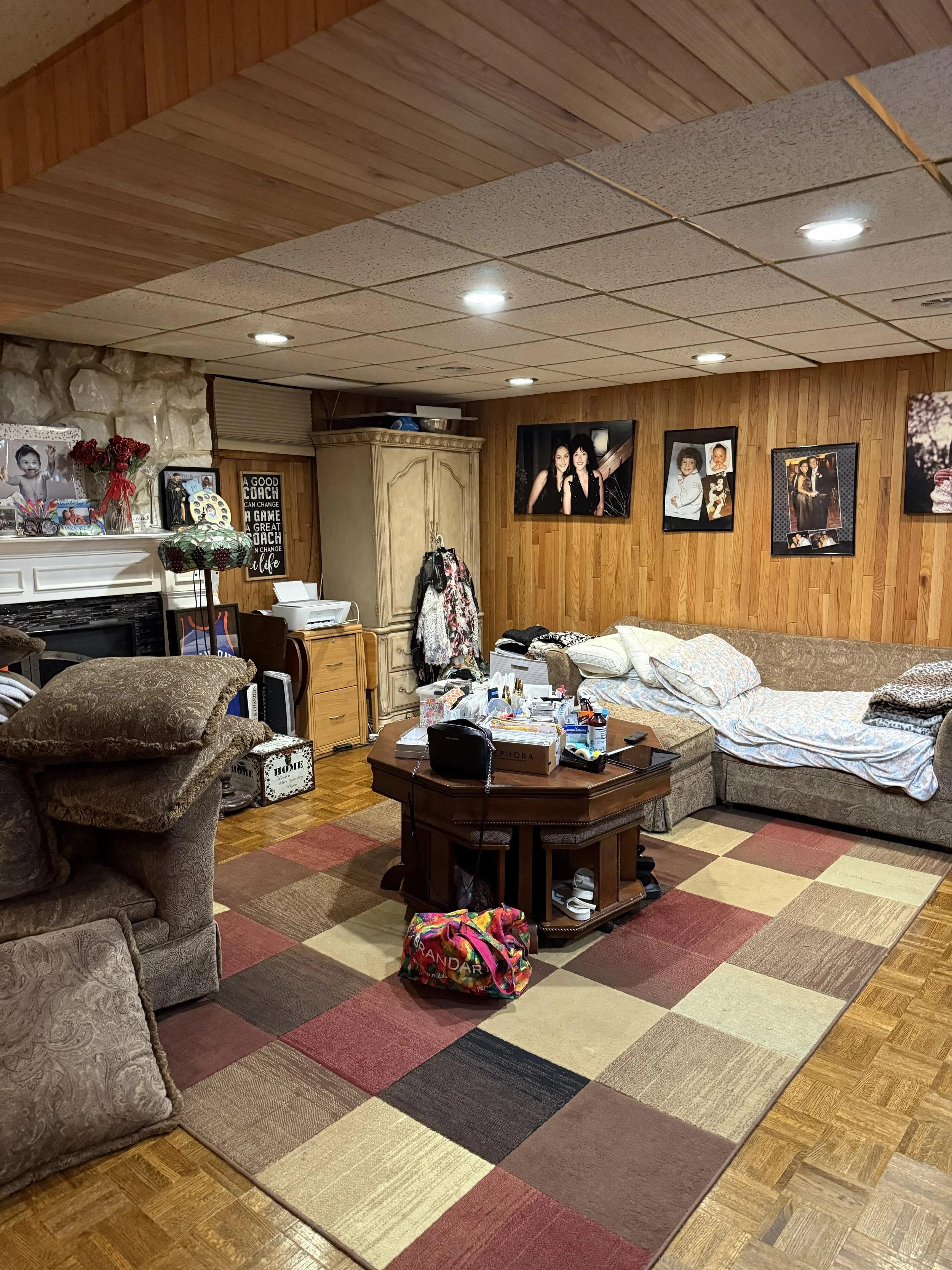 A cozy living room with wooden-paneled walls and ceiling, decorated with family photos and artwork. There are comfortable couches, a table cluttered with various items, a multicolored area rug on the hardwood floor, and a lamp with a stained glass shade.