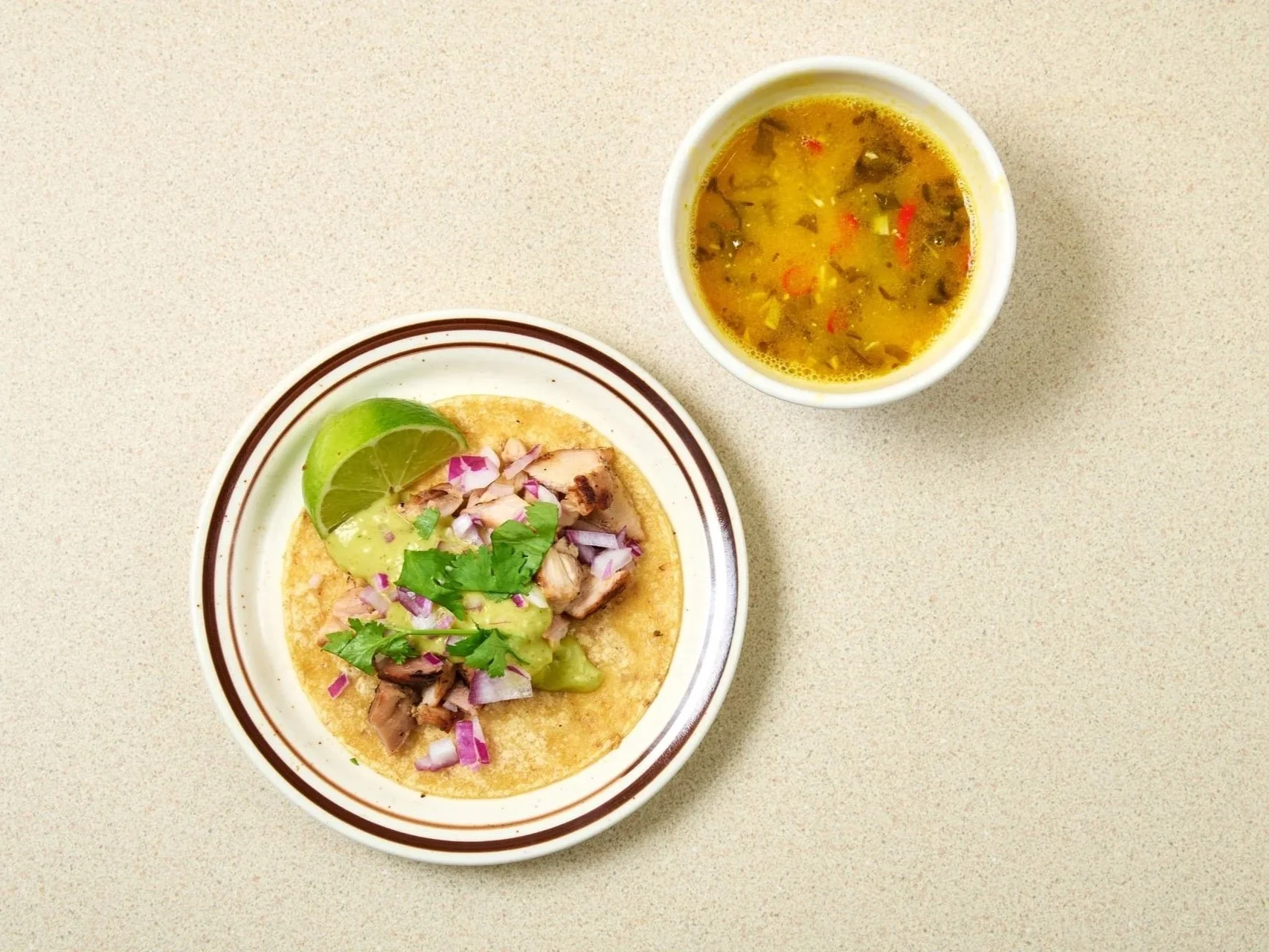 A bowl of chicken tostada topped with avocado, red onion, cilantro, and a wedge of lime, accompanied by a cup of chicken soup with vegetables on a beige countertop.