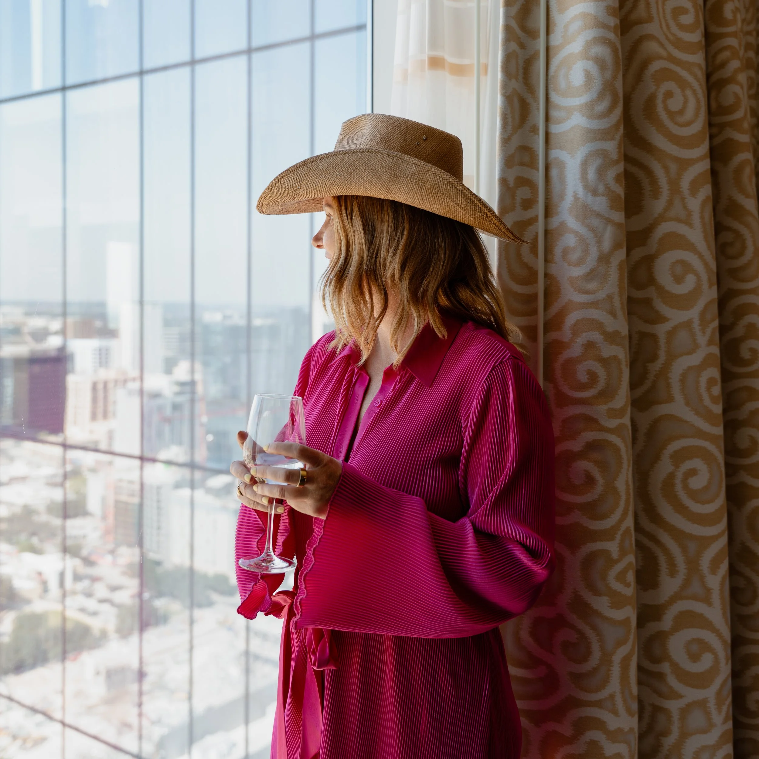 A woman in a pink long-sleeve shirt and a wide-brimmed cowboy hat holding a wine glass, standing near a large window in a high-rise building with a city view.