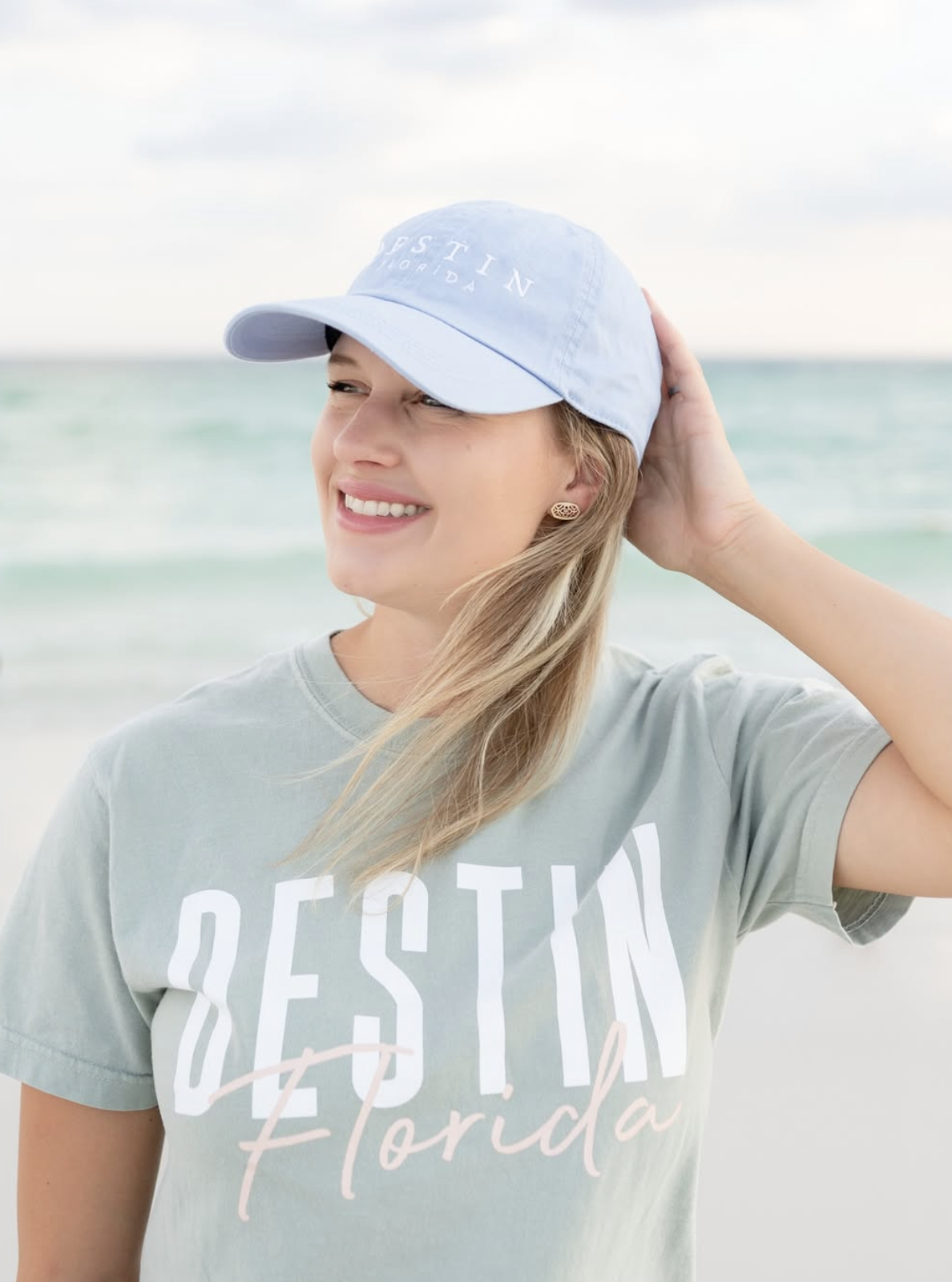 Smiling woman on the beach wearing a light blue cap and a grey t-shirt with 'DESTIN Florida' written on it.