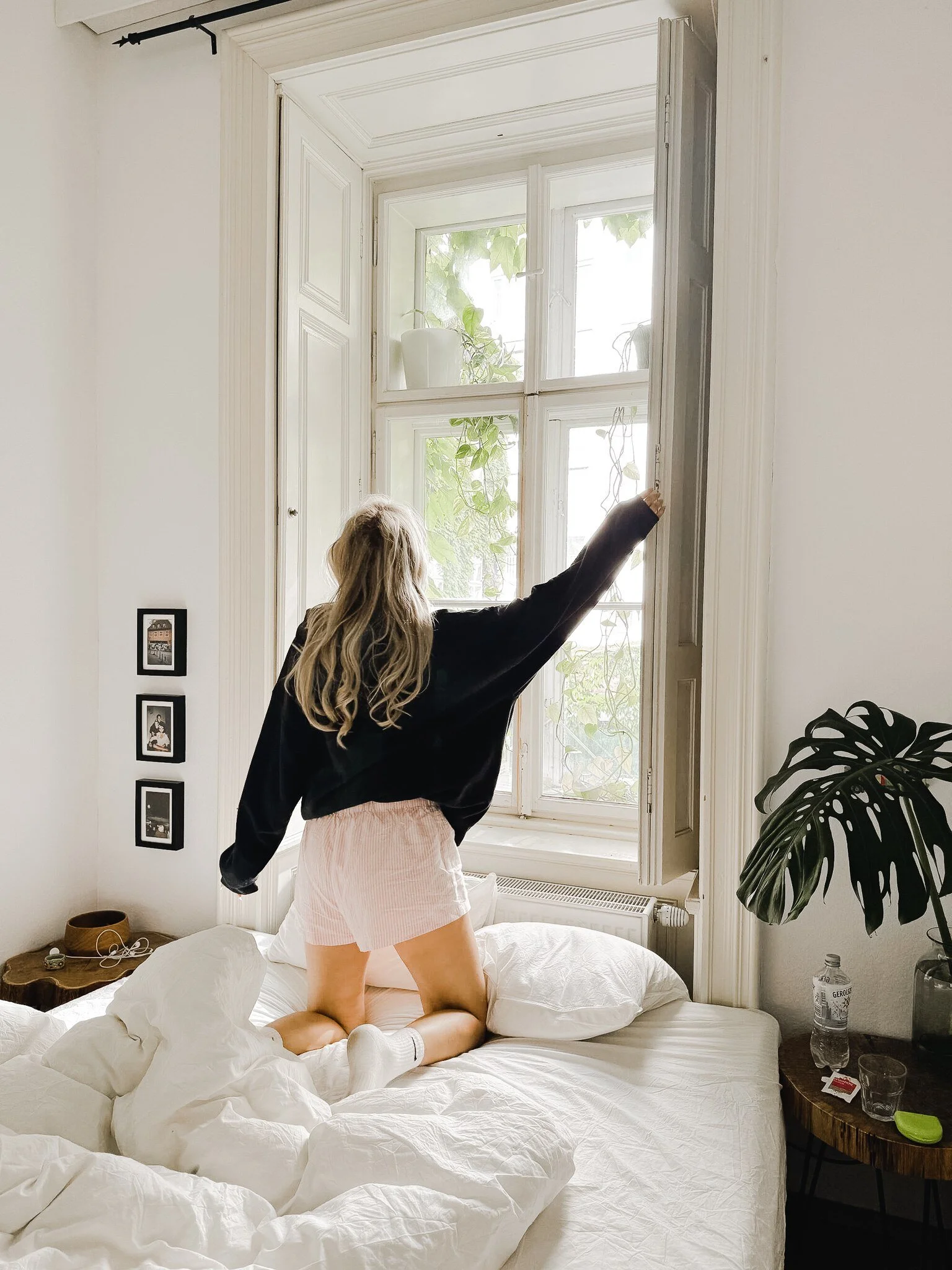 A woman kneeling on a bed, reaching towards a large window, with bean bag pillows and a water bottle on a side table in a bright, white room.