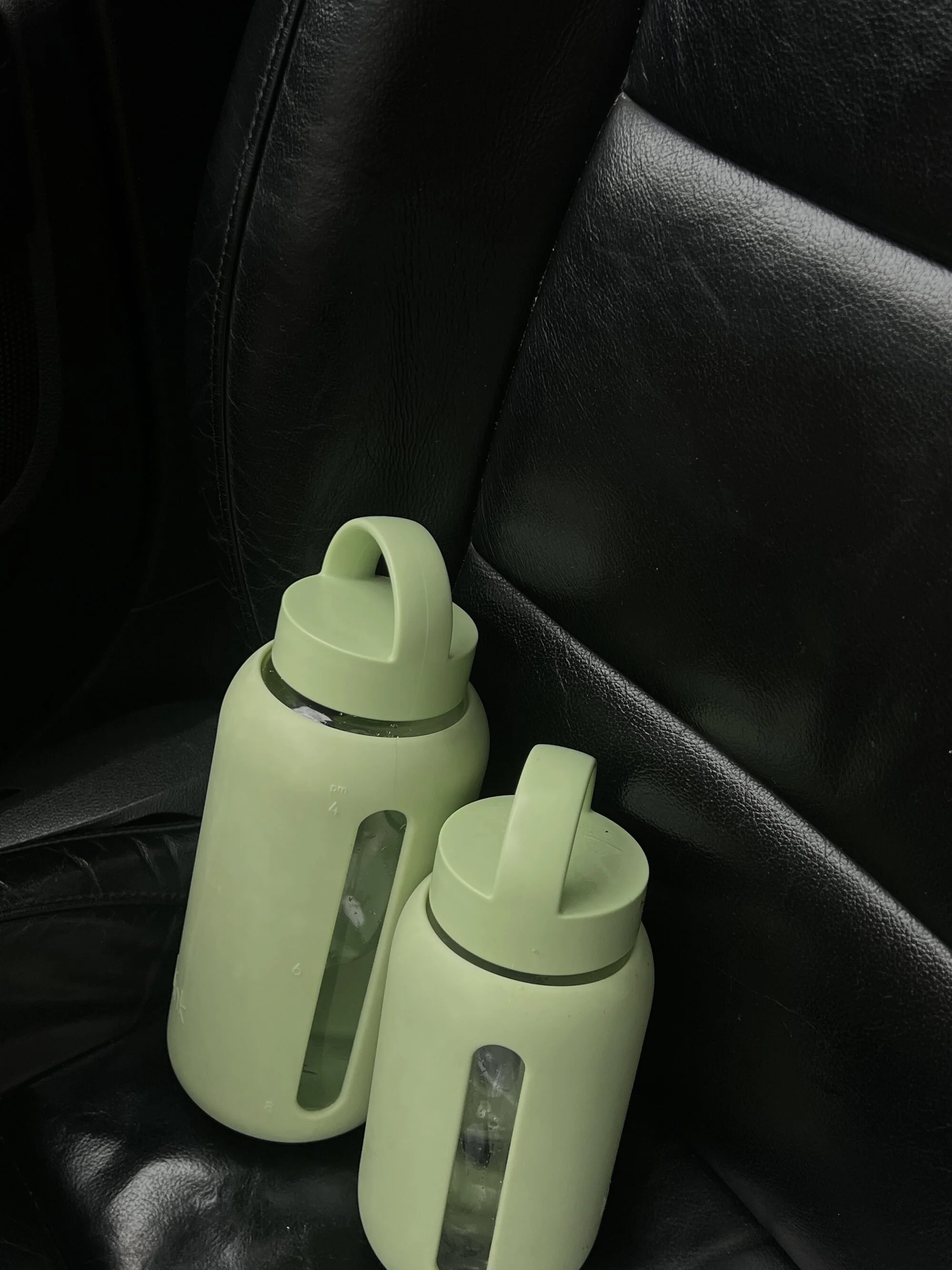 Two light green water bottles with handles and transparent water measurement windows, resting on a black leather car seat.